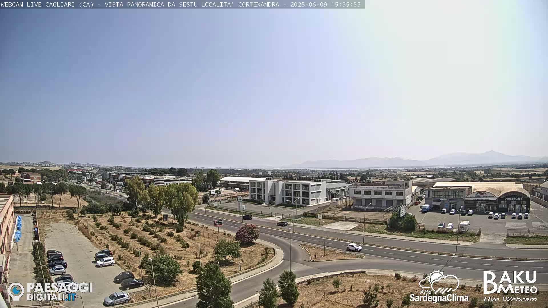 A panoramic view of a city with industrial buildings and a road intersection under a clear, sunny sky, with distant mountains visible on the horizon.