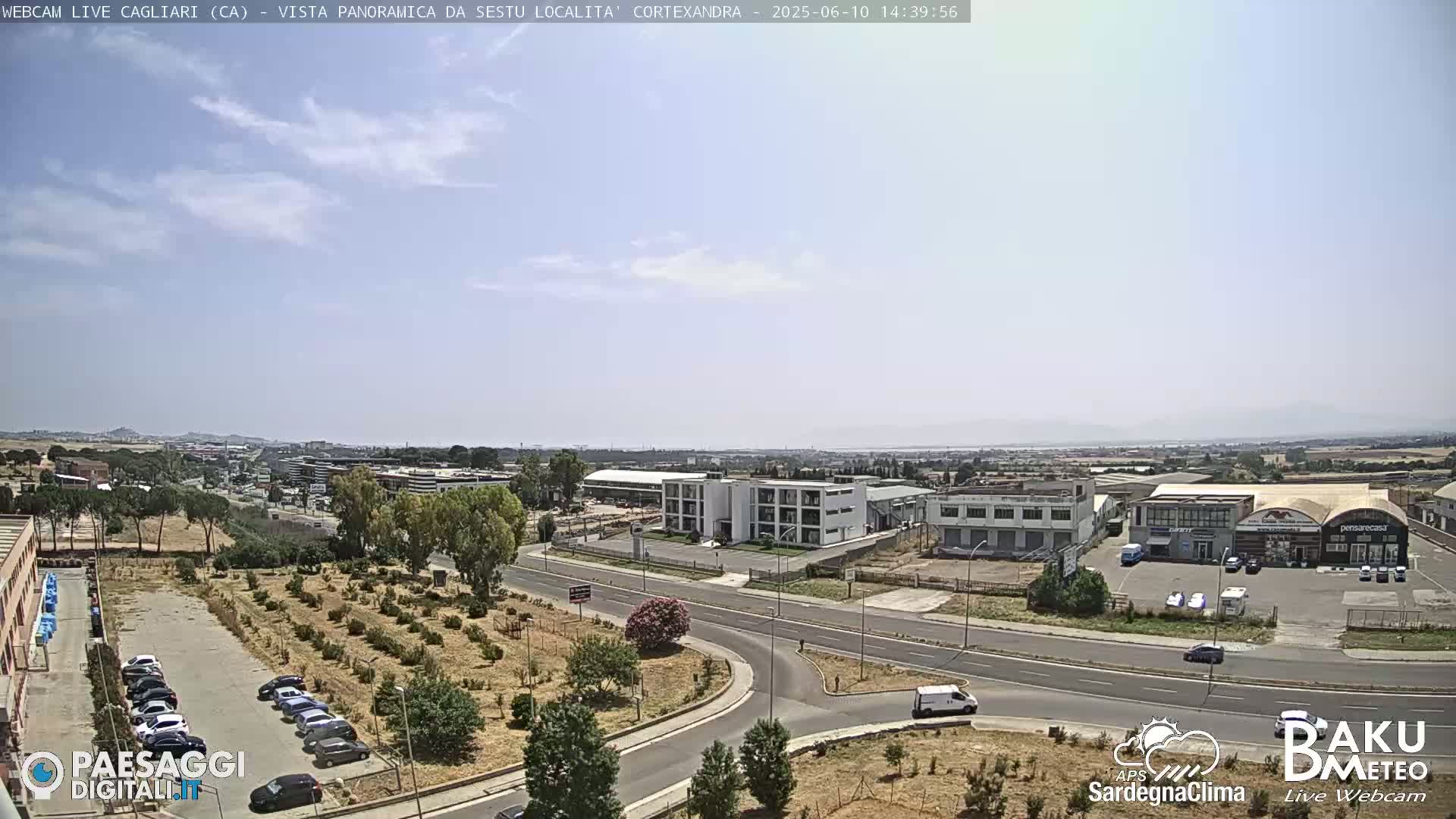 A sunny, mostly clear day reveals a wide view of a city's outskirts, featuring roads, industrial buildings, and sparse vegetation.