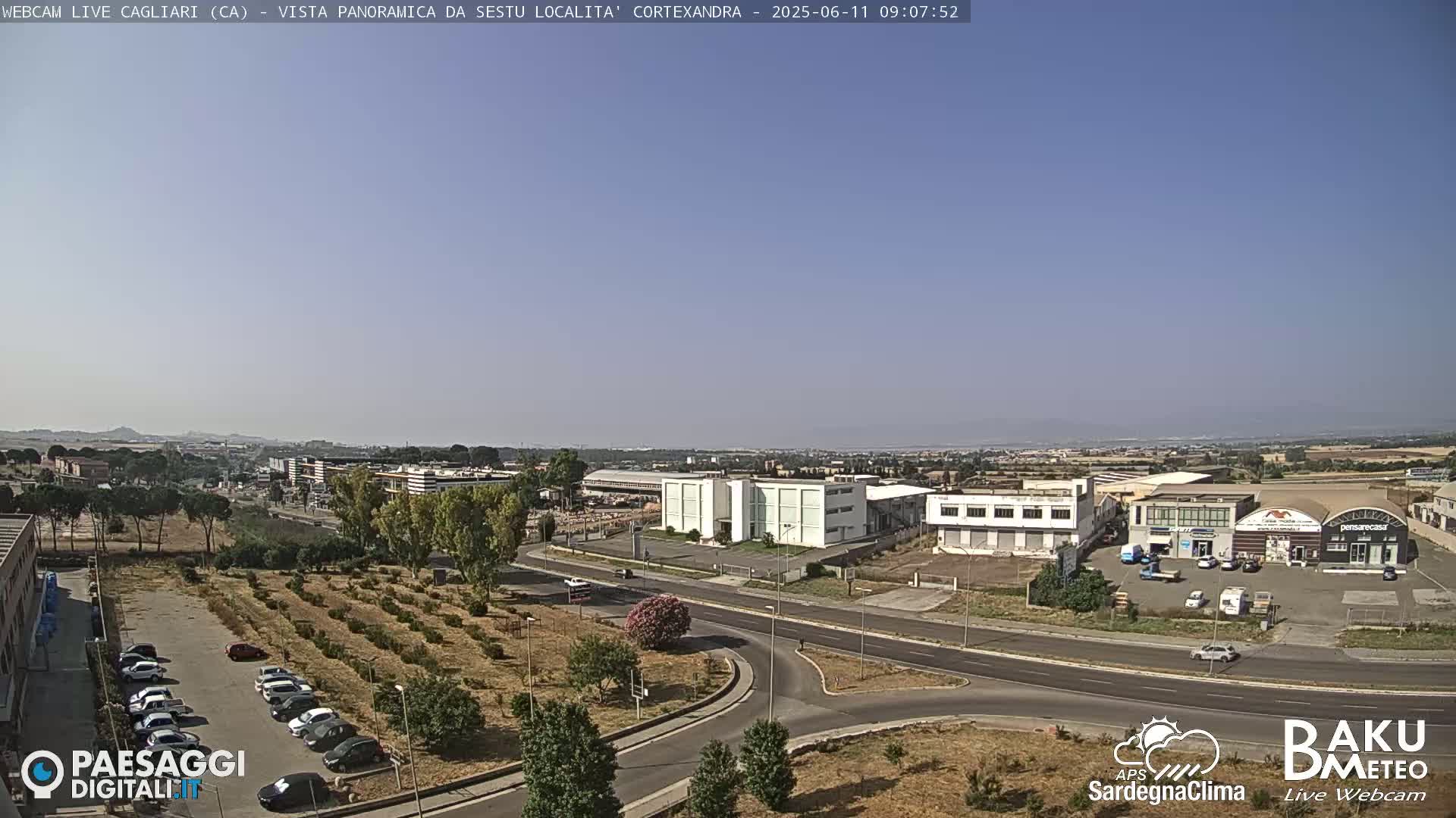 A sunny, clear day reveals a panoramic view of a city's outskirts, featuring industrial buildings, roads, and a parking lot filled with cars, alongside a field of low-growing plants.