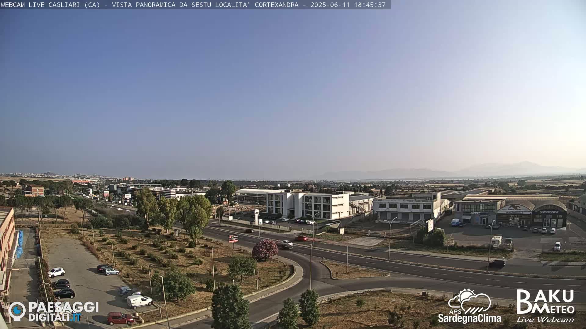 A clear, sunny day reveals a panoramic view of a suburban area with low-rise buildings, roads, and sparse vegetation, mountains visible in the distance.