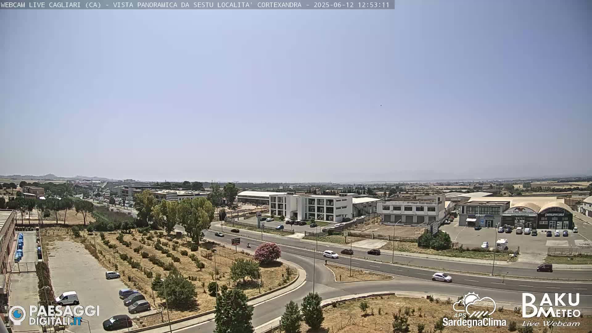A panoramic view of a city under a clear, sunny sky shows roads, buildings, and sparse vegetation.