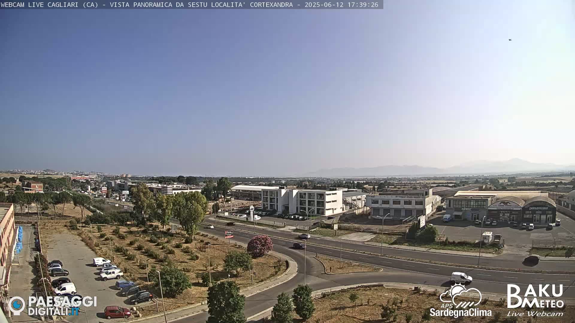 A panoramic view of a city under a clear, sunny sky shows roads, buildings, and sparse vegetation.