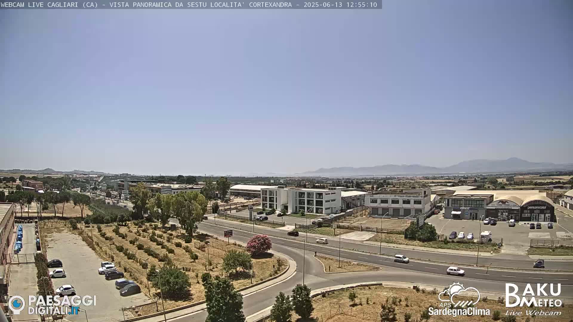 A clear, sunny day reveals a wide shot of an industrial area and surrounding landscape, including roads, buildings, and some vegetation.