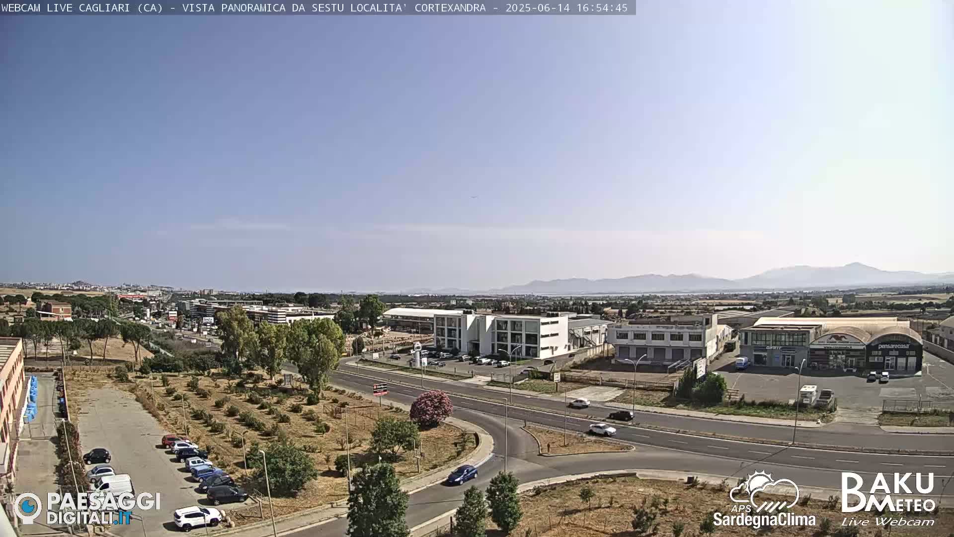 A sunny, clear day reveals a panoramic view of an industrial area with buildings, roads, and parked cars, stretching towards distant mountains under a pale blue sky.