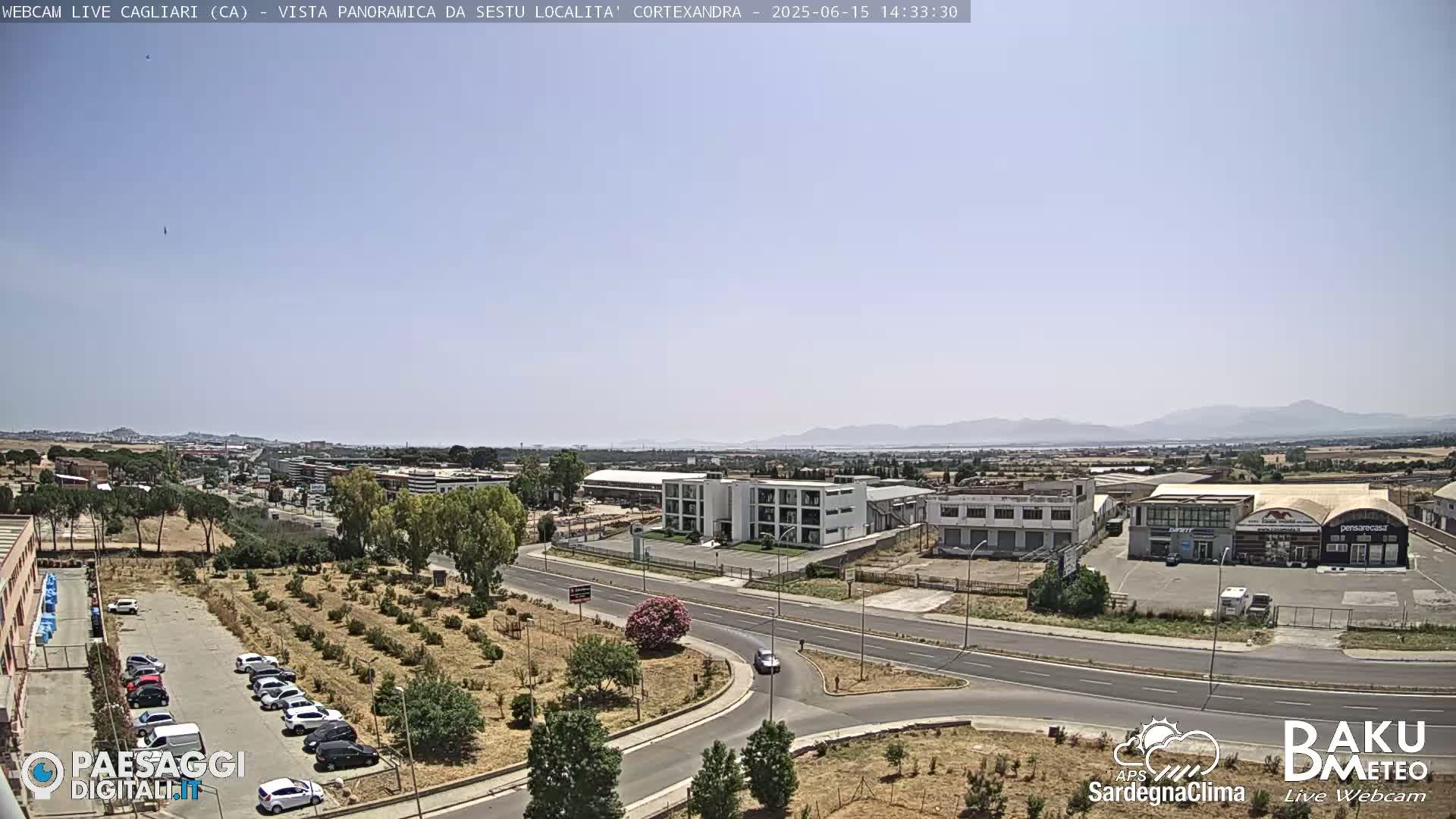 A clear, sunny day reveals a wide shot of a road intersection, industrial buildings, and a parking lot surrounded by sparse vegetation, with mountains visible in the distance.