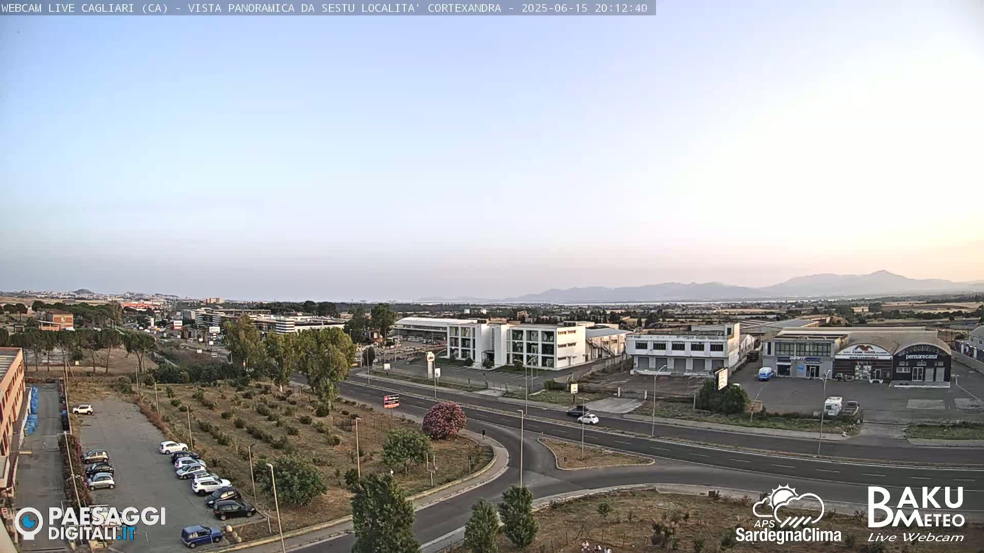 A panoramic view of a city under a clear, light-blue sky shows a road system intersecting with a parking lot containing several cars, and numerous buildings, including what appears to be a commercial center,  with distant mountains visible on the horizon.