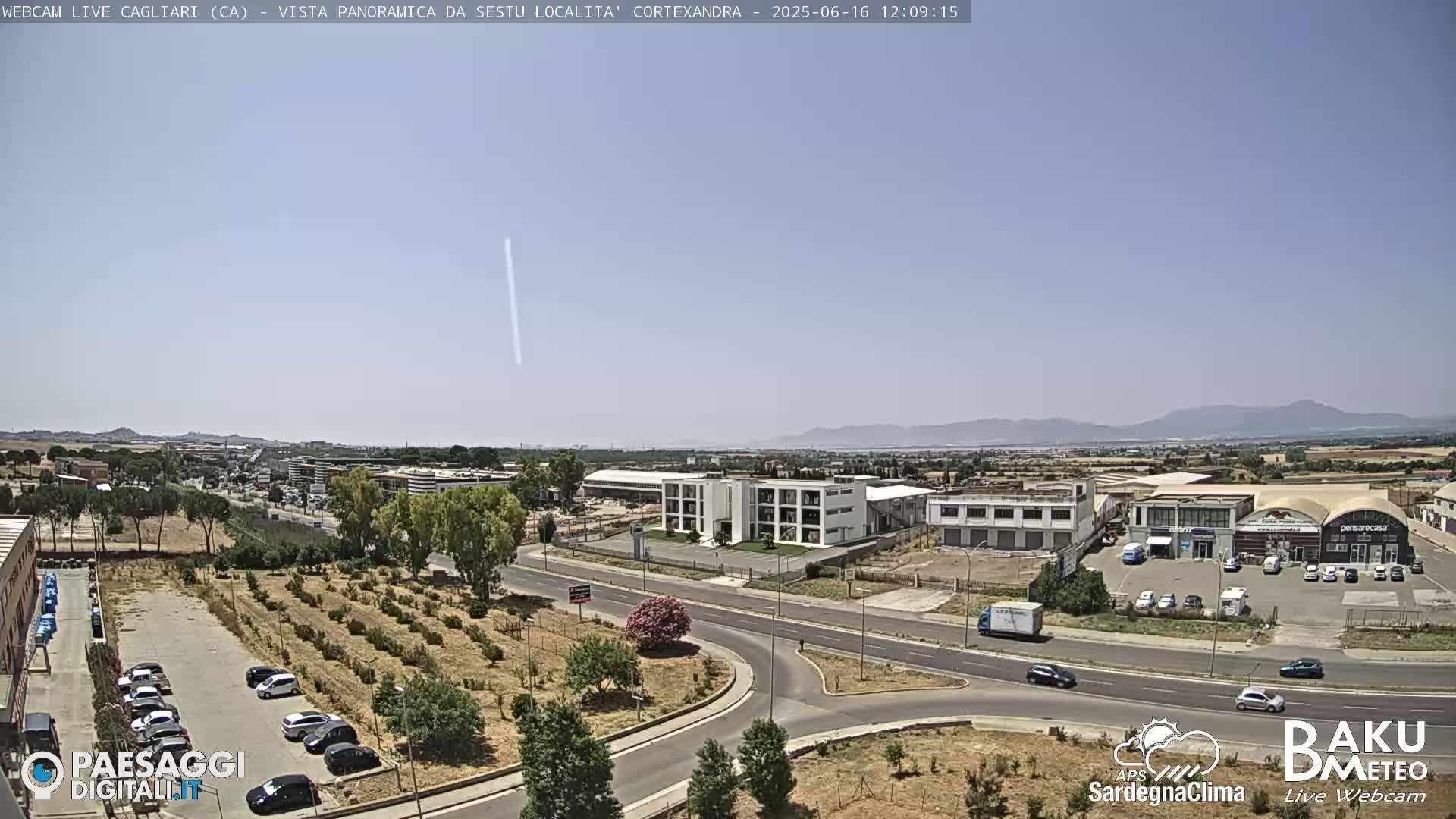A sunny, clear day reveals a wide shot of a roadway intersection and industrial area, surrounded by sparse vegetation and distant mountains.