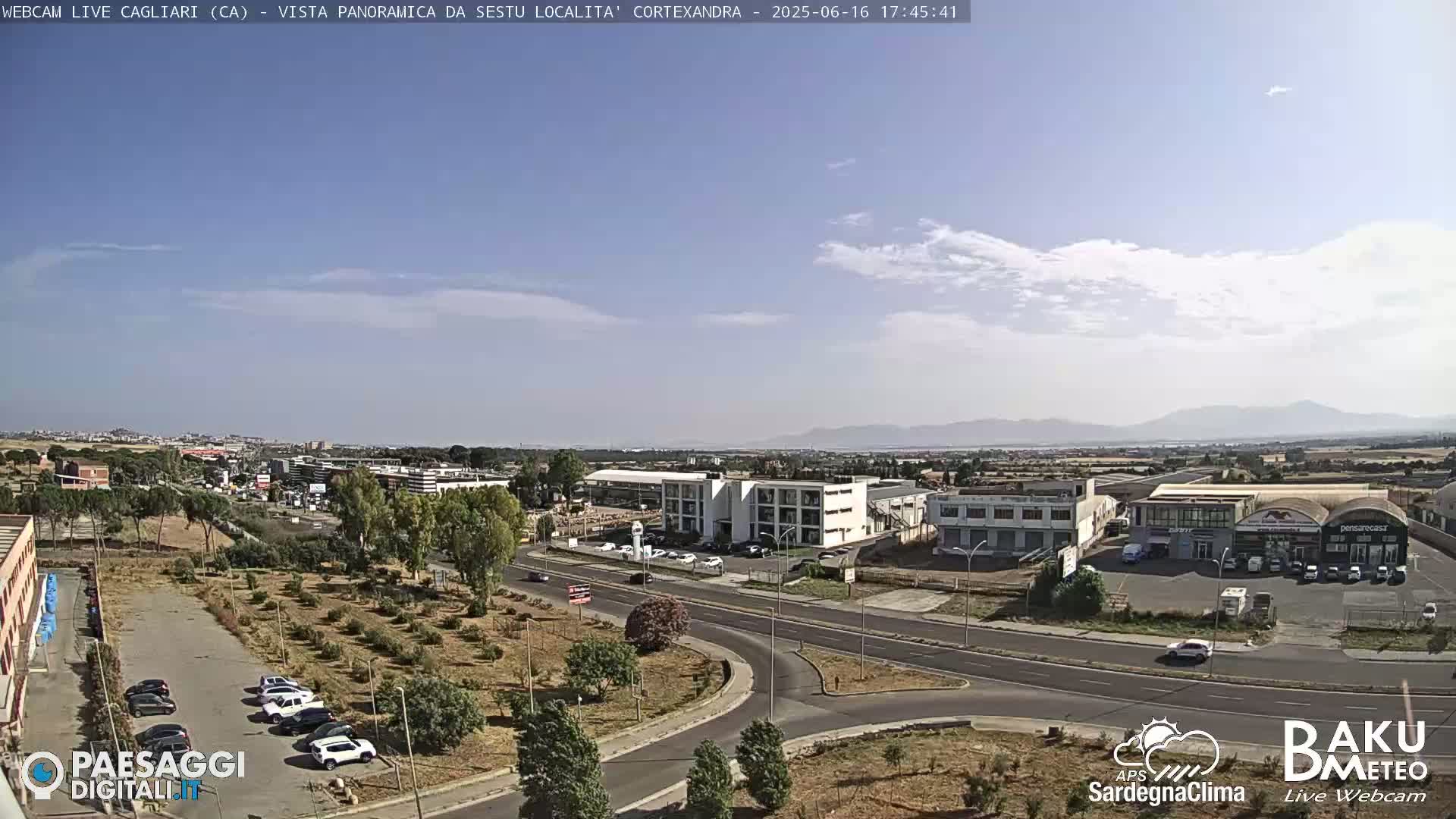A mostly sunny day reveals a panoramic view of a city with a highway, buildings, and a parking lot containing several cars, all situated against a backdrop of distant mountains.