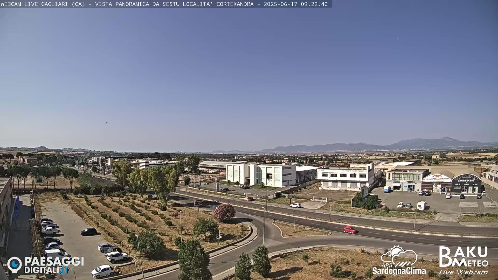 A panoramic view of a city under a clear blue sky shows a road intersection near industrial buildings and a cultivated field.