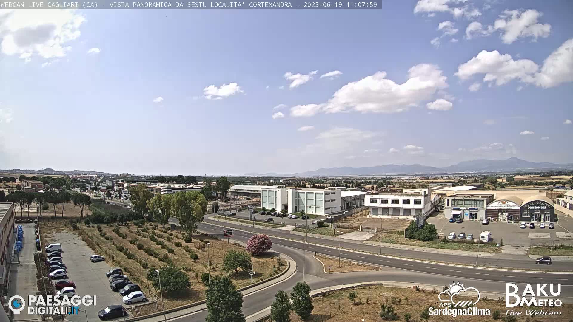 A partly cloudy day reveals a wide shot of a roadway intersection, surrounded by industrial buildings and a sparse, scrubby landscape, with distant mountains under a light blue sky.