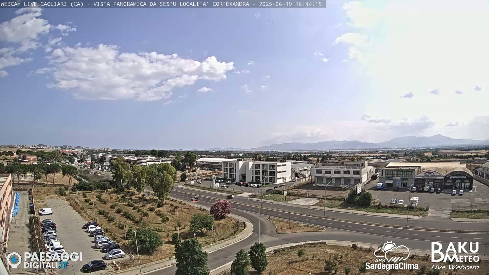 A sunny day with scattered clouds reveals a panoramic view of an industrial area, parking lots, and roadways, with mountains visible in the distance.