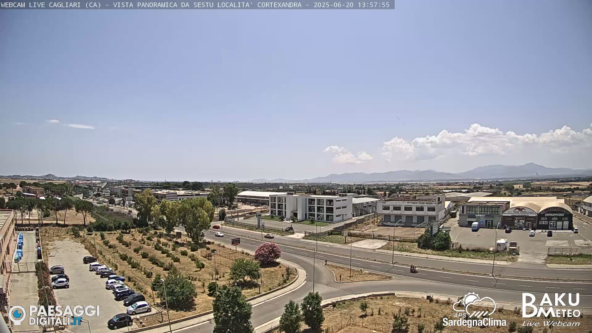 A high-angle view of a sunny road intersection surrounded by industrial buildings and sparse vegetation, with a mountain range visible in the distance under a mostly clear sky with some cumulus clouds.