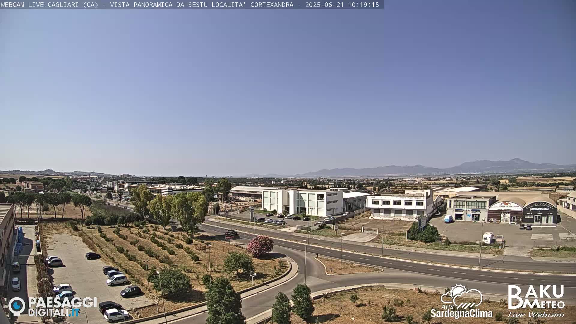 A clear, sunny day reveals a panoramic view of a suburban area with industrial buildings, roads, and sparse vegetation, mountains visible in the distance.