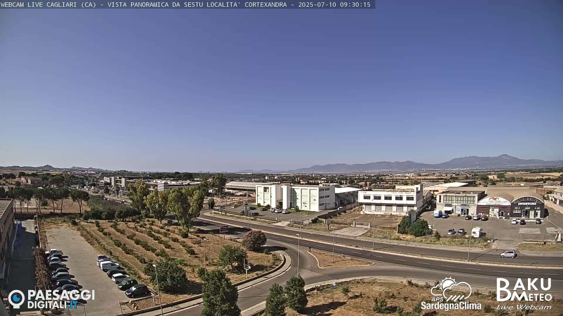 A clear, sunny day overlooks a suburban area with industrial buildings, roads, and a parking lot filled with cars, all set against a backdrop of distant mountains.