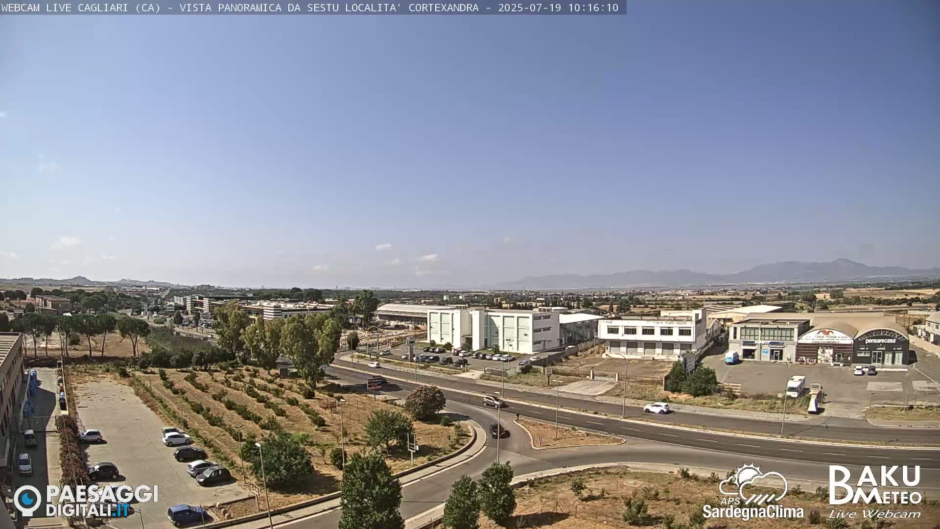 A panoramic view of a city under a clear, sunny sky, showing roads, buildings, and a small orchard.