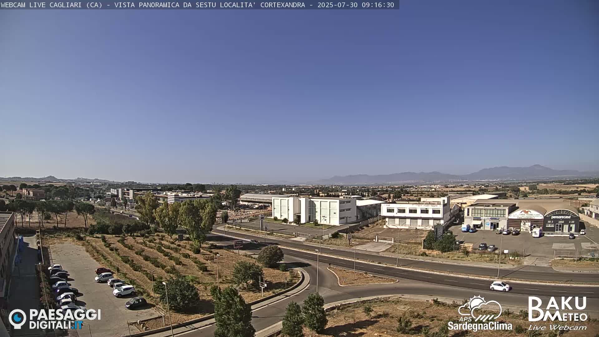 A clear, sunny day reveals a panoramic view of a suburban area with industrial buildings, roads, and sparse vegetation, extending to distant mountains under a clear blue sky.