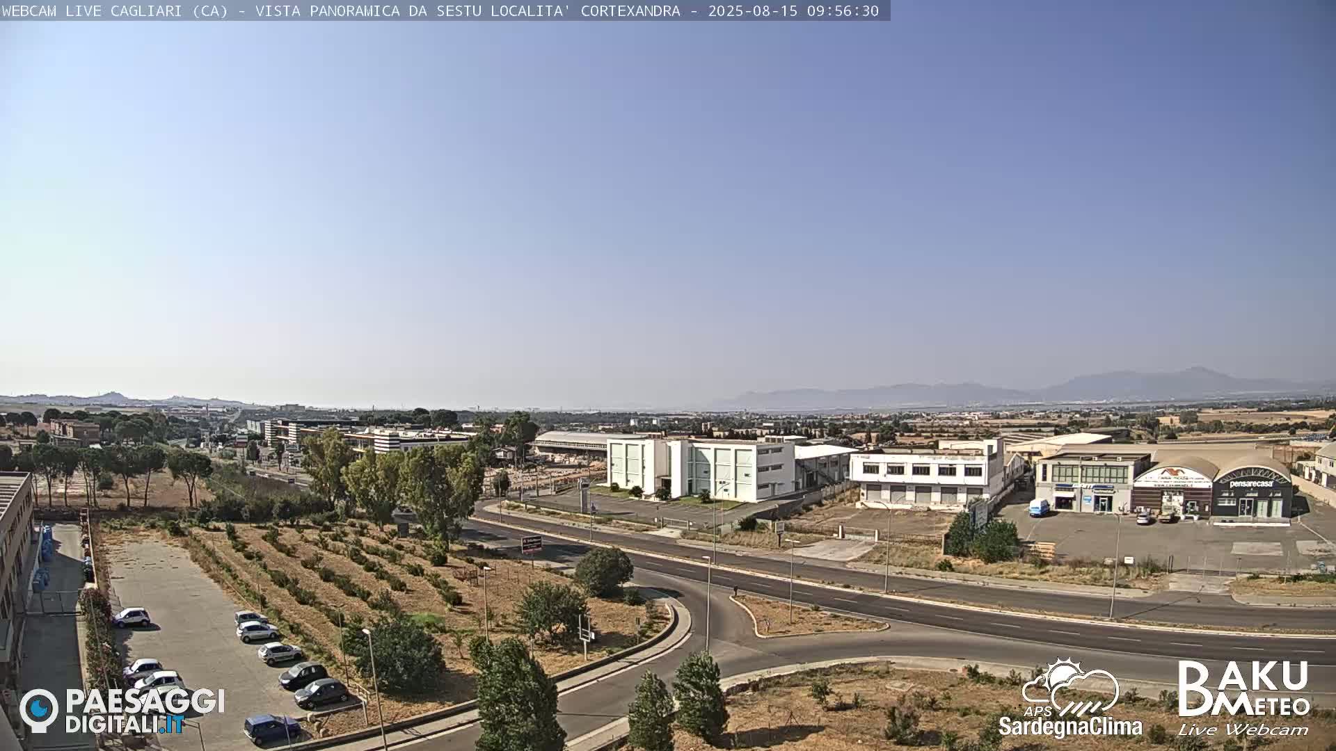 A panoramic view on a clear, sunny day shows a road system intersecting with industrial buildings, businesses, and a sparse parking lot next to some small trees and shrubs; distant mountains are visible in the background.