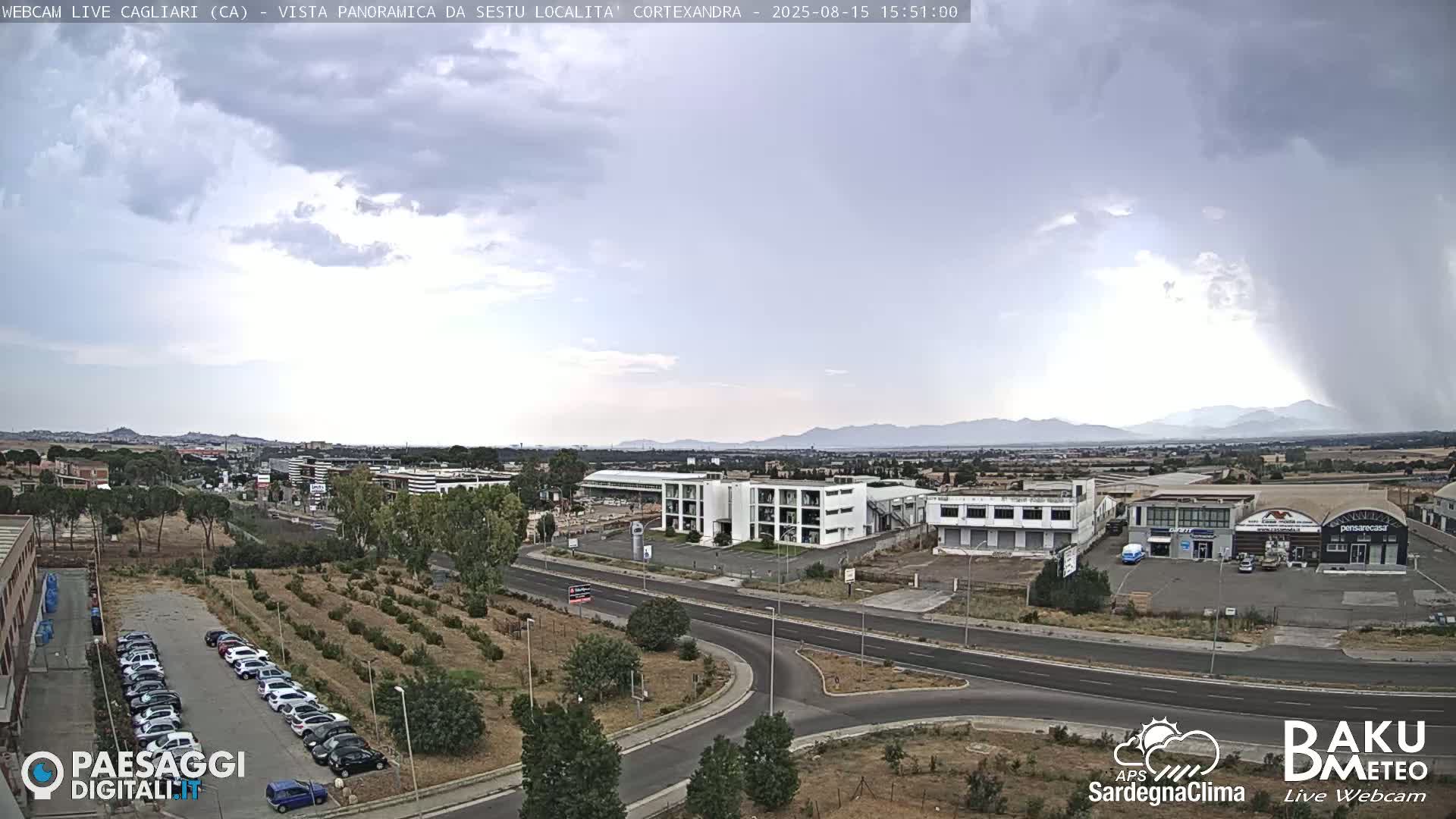 A panoramic view on a clear, sunny day shows a road system intersecting with industrial buildings, businesses, and a sparse parking lot next to some small trees and shrubs; distant mountains are visible in the background.