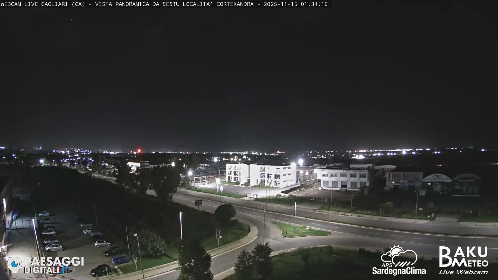 The image displays a nighttime panoramic view of a city with illuminated roads, several buildings, a parking lot with cars, and distant urban lights under a dark, clear sky.