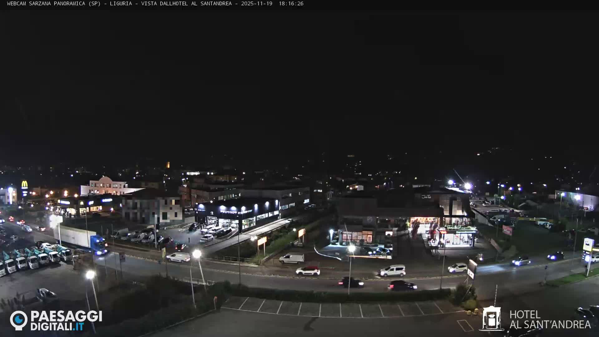 The image displays a nighttime panoramic view of a city with illuminated roads, several buildings, a parking lot with cars, and distant urban lights under a dark, clear sky.