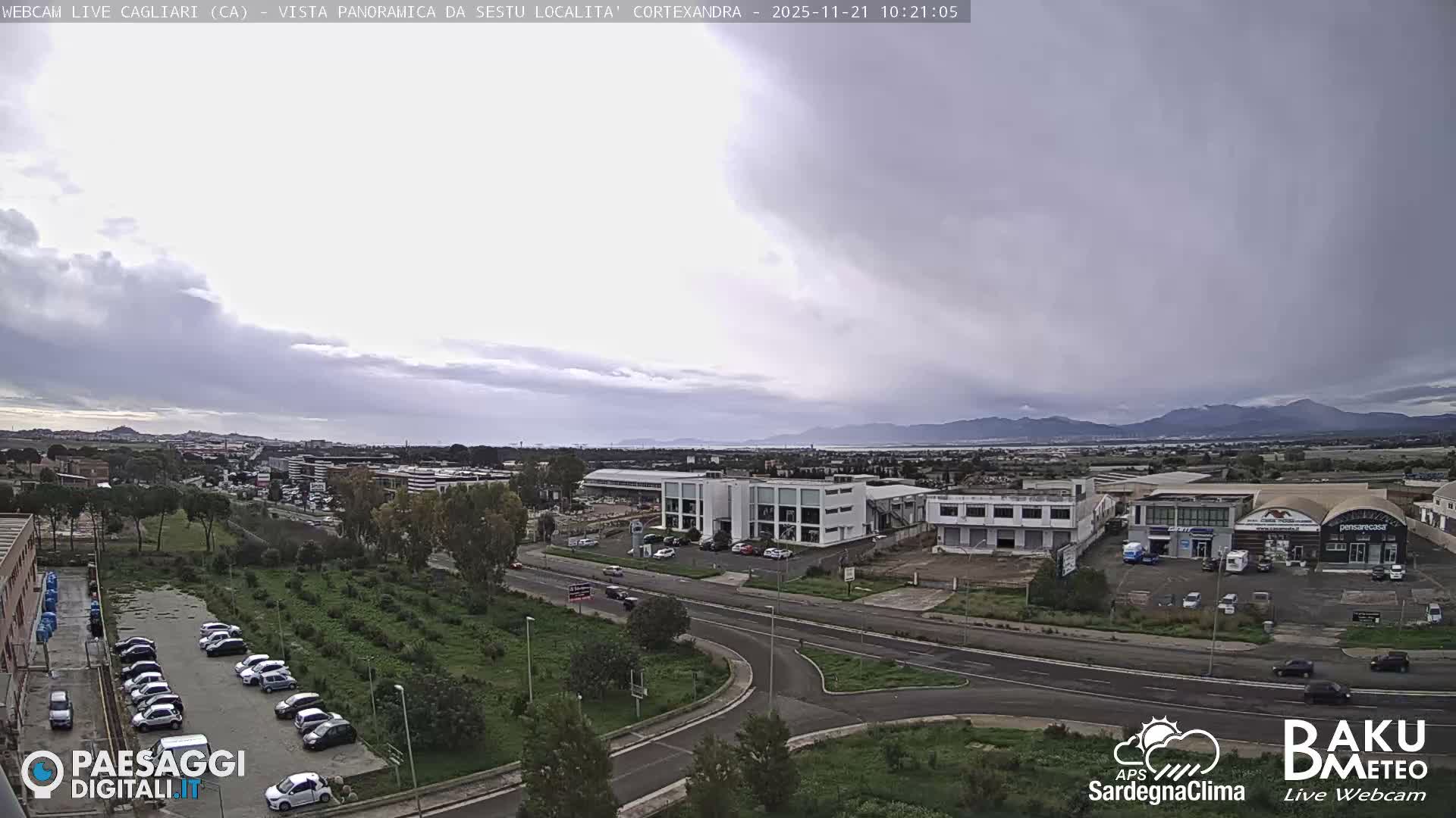 The image depicts an urban-industrial landscape under an overcast and cloudy sky, featuring multiple buildings, busy roads with cars, several parking lots, and distant hills.