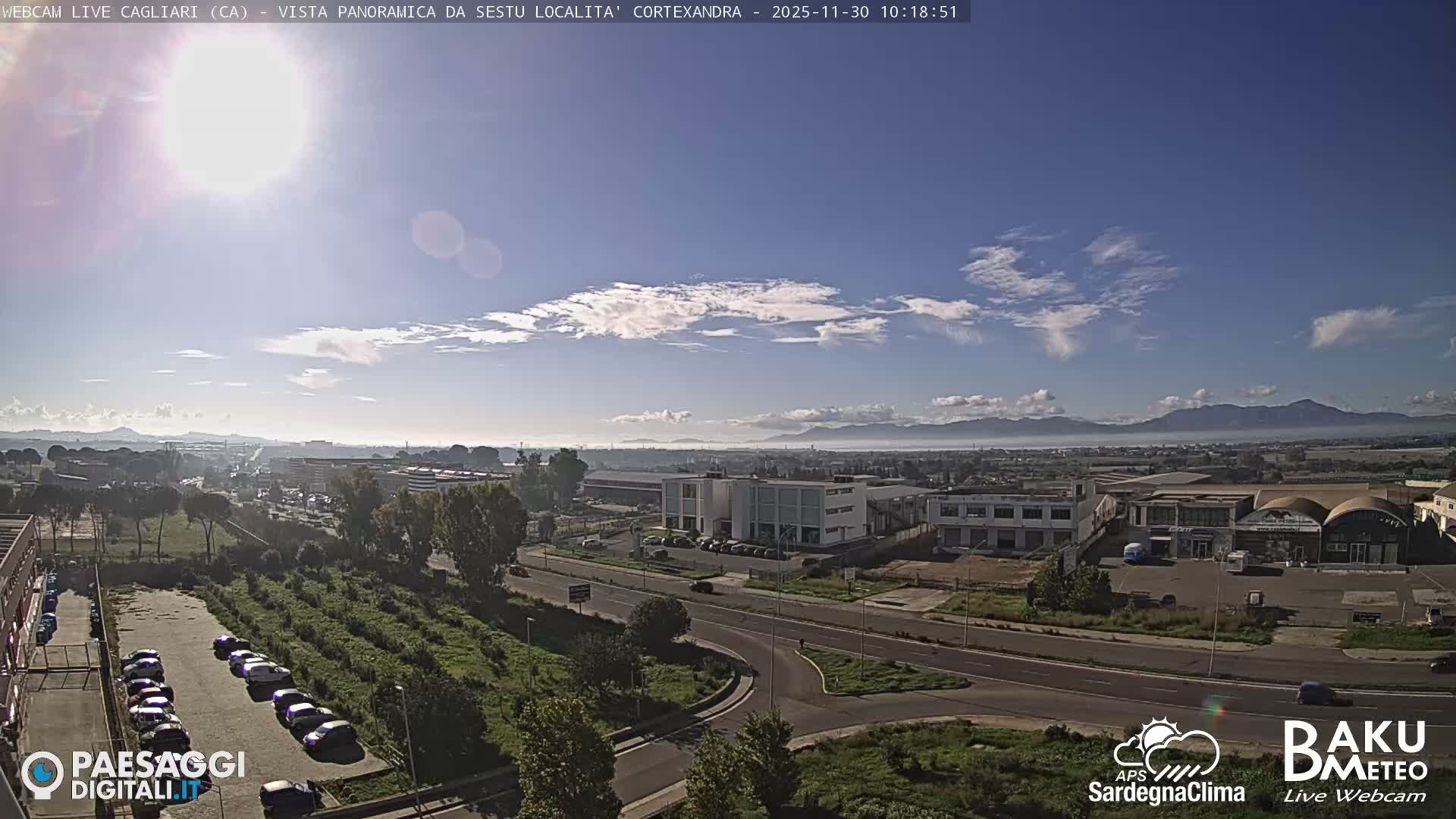 The image shows a sunny, clear day with scattered white clouds over a sprawling urban landscape featuring roads, buildings, parked cars, and distant hazy mountains under a bright blue sky.