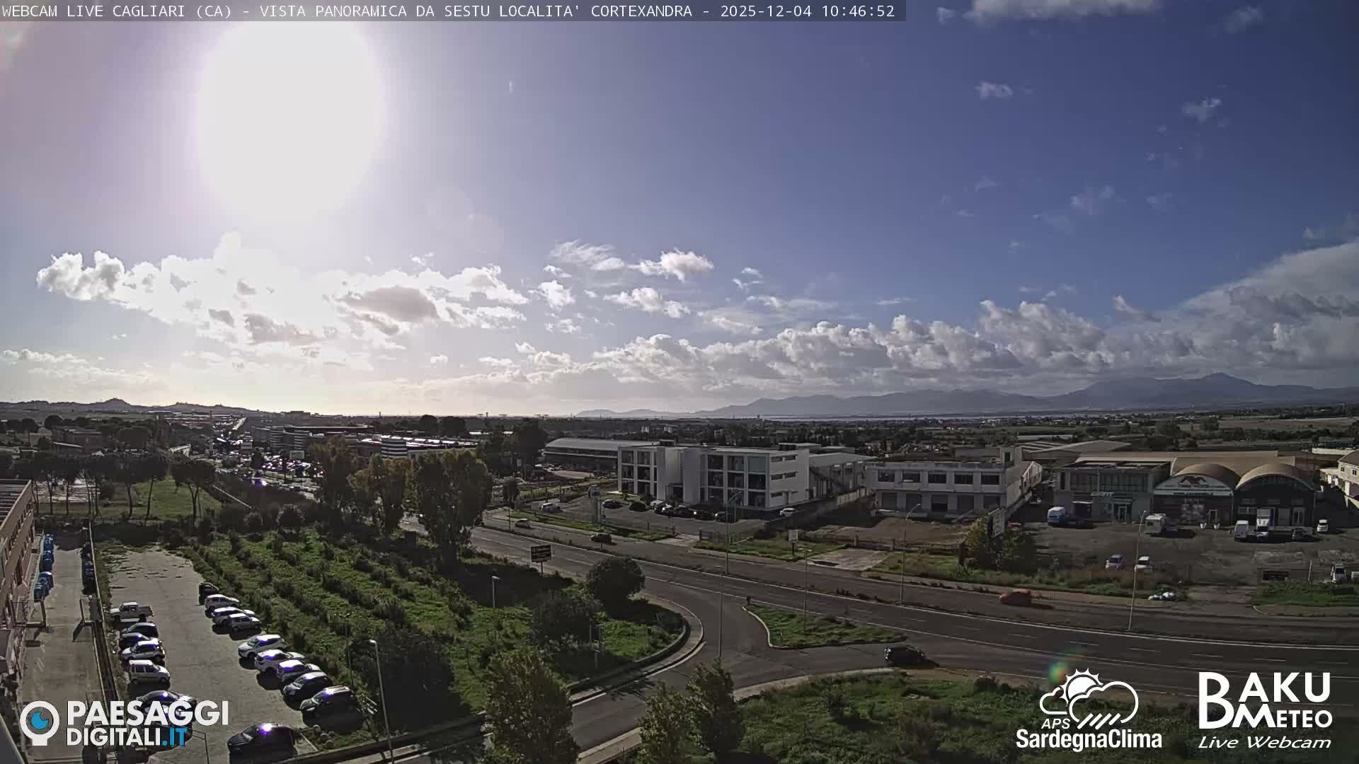 An elevated view reveals a sprawling semi-urban landscape with numerous buildings, roads, and parking lots, flanked by green spaces and distant mountains, all under a bright, partly cloudy sky.