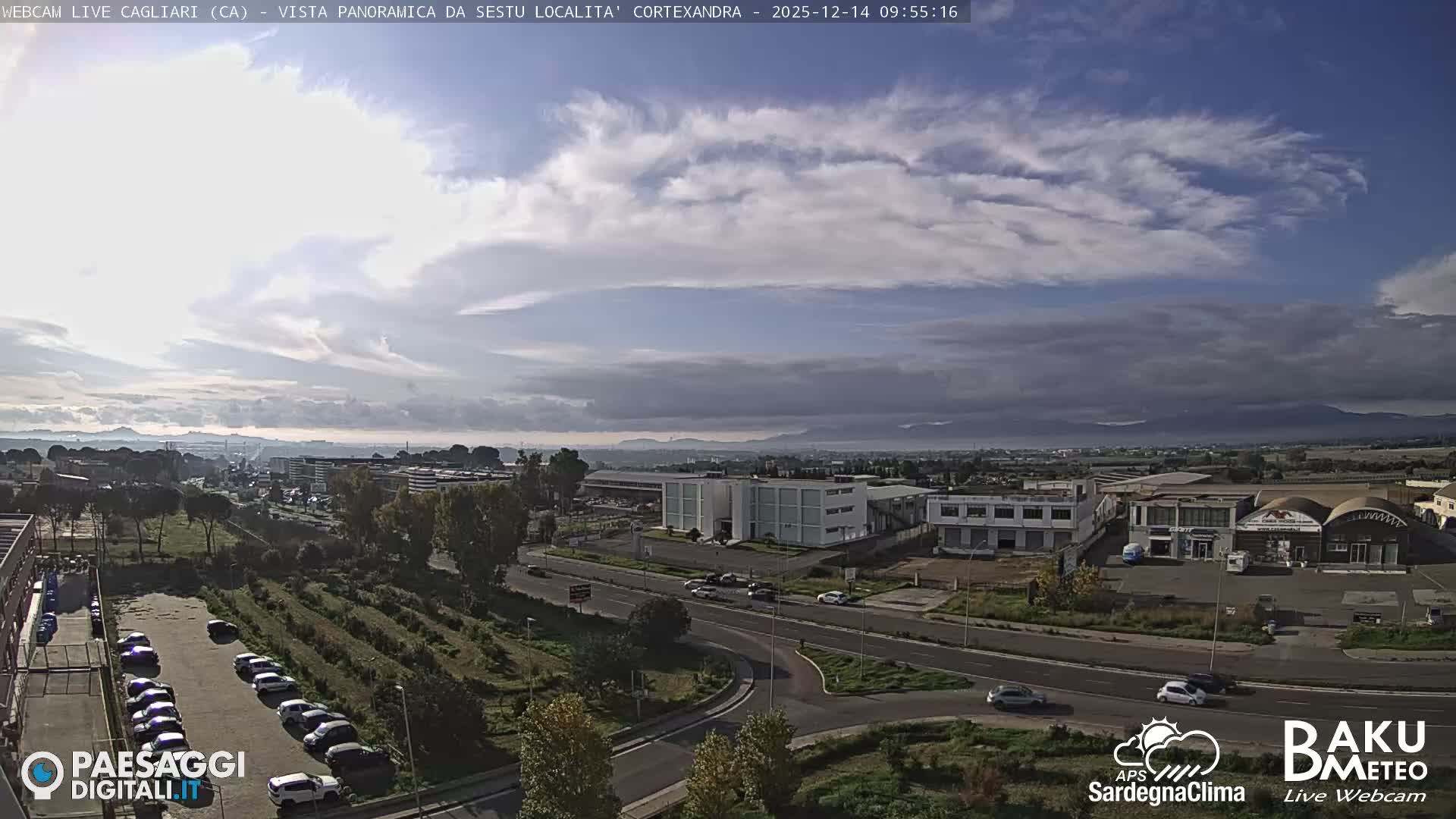 An elevated view reveals a sprawling semi-urban landscape with numerous buildings, roads, and parking lots, flanked by green spaces and distant mountains, all under a bright, partly cloudy sky.