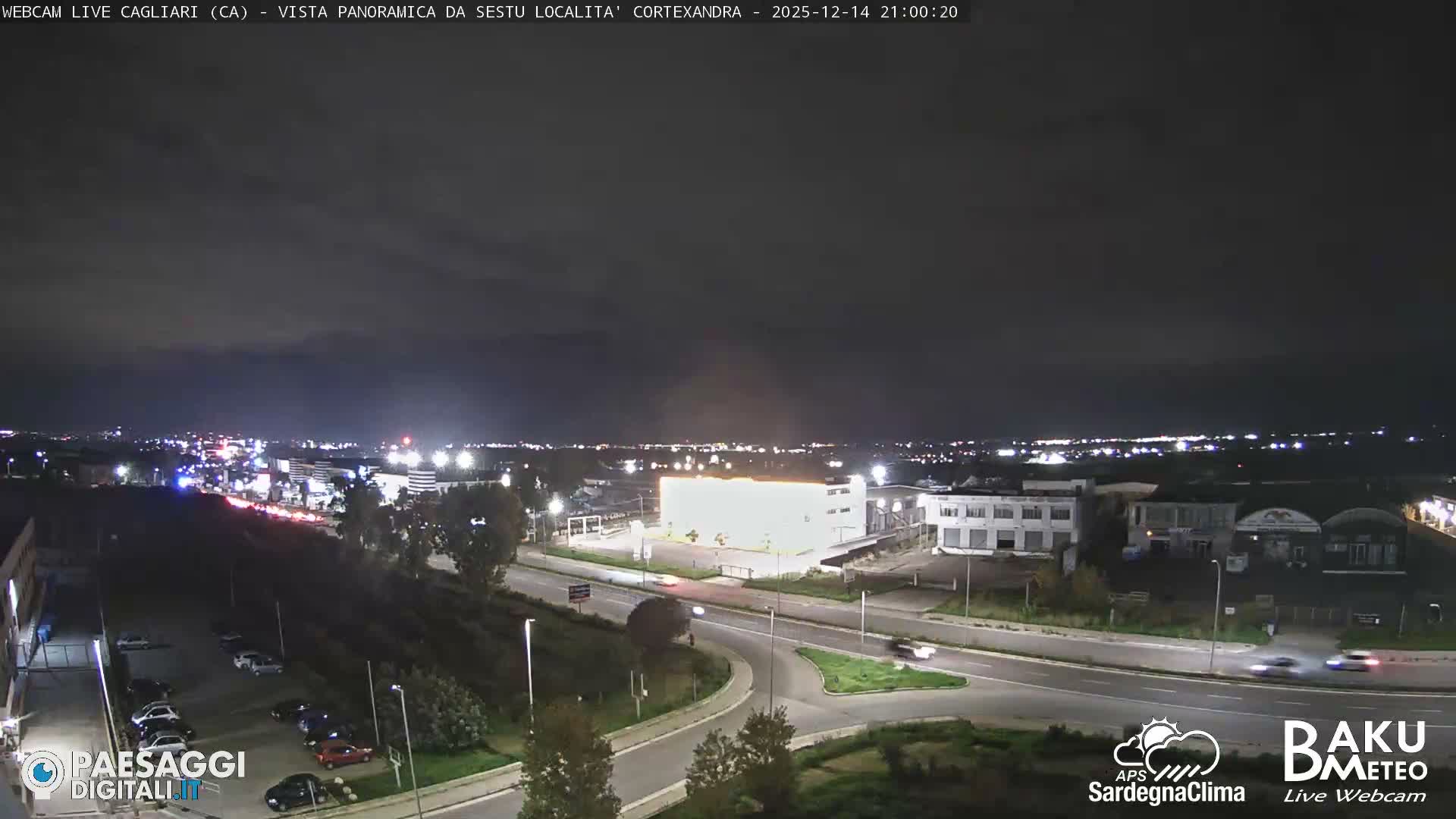 An elevated view reveals a sprawling semi-urban landscape with numerous buildings, roads, and parking lots, flanked by green spaces and distant mountains, all under a bright, partly cloudy sky.