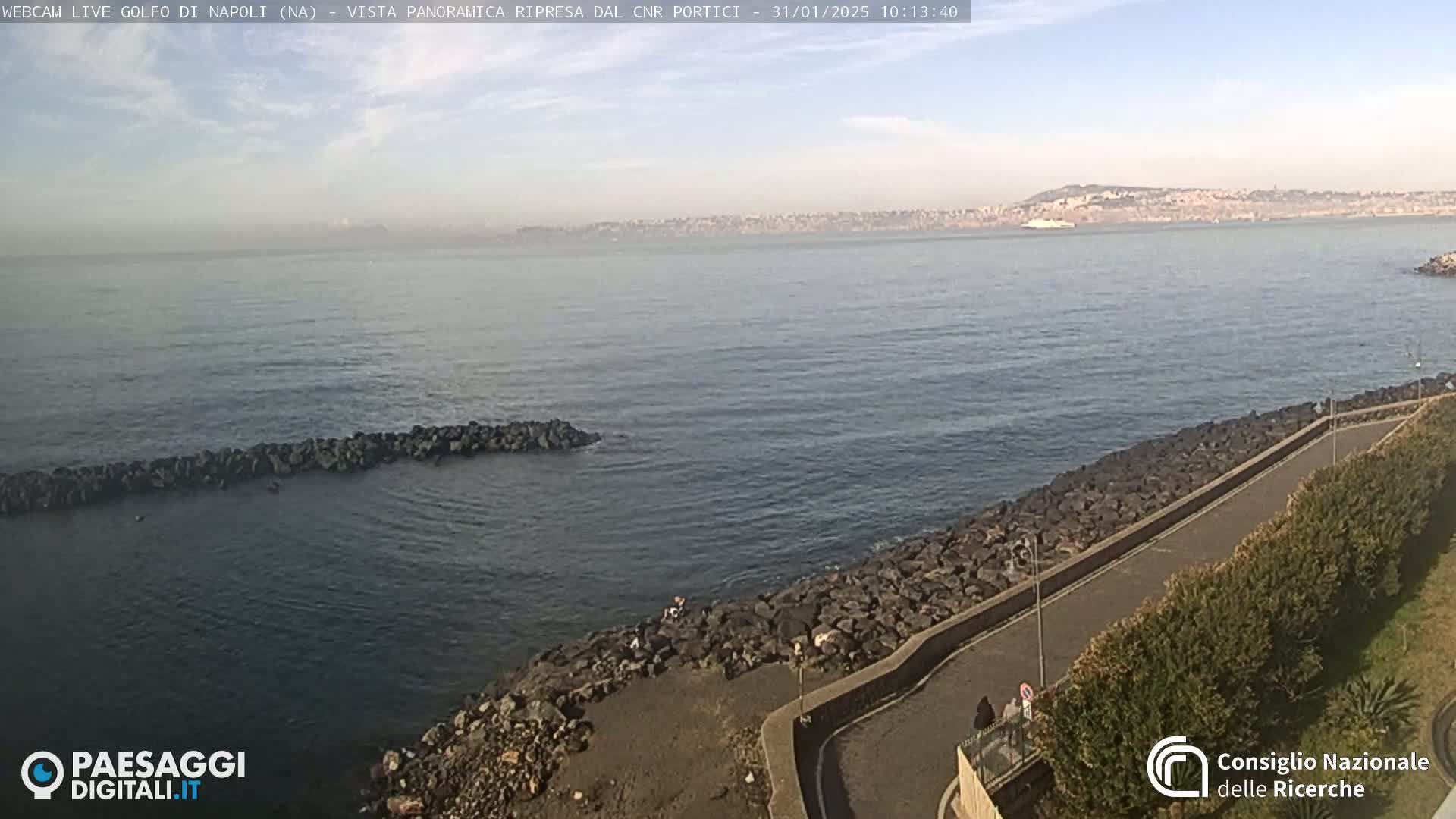 A rocky shoreline curves gently around a calm bay, with a distant city skyline visible under a partly cloudy sky.