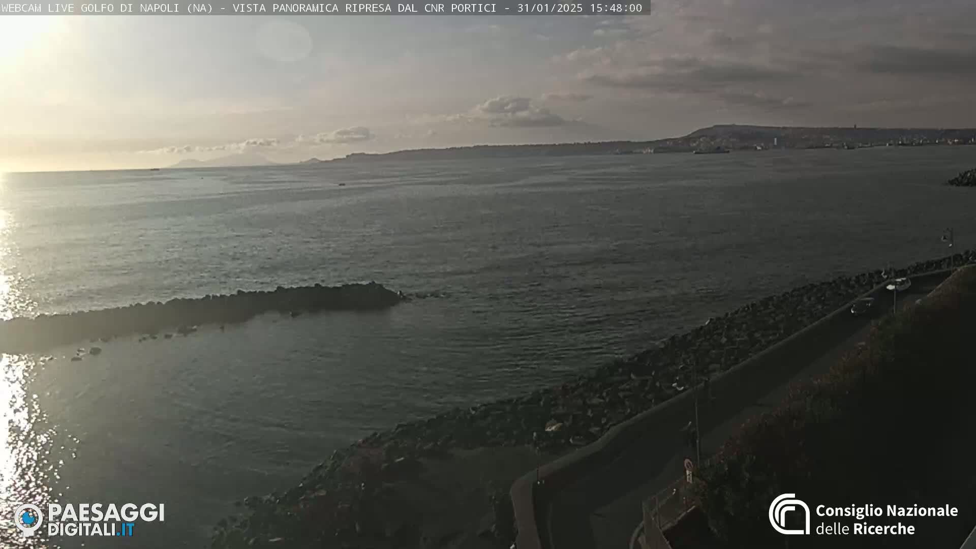 A panoramic view of a calm sea, under a partly cloudy sky, shows a rocky shoreline with a road running along it.