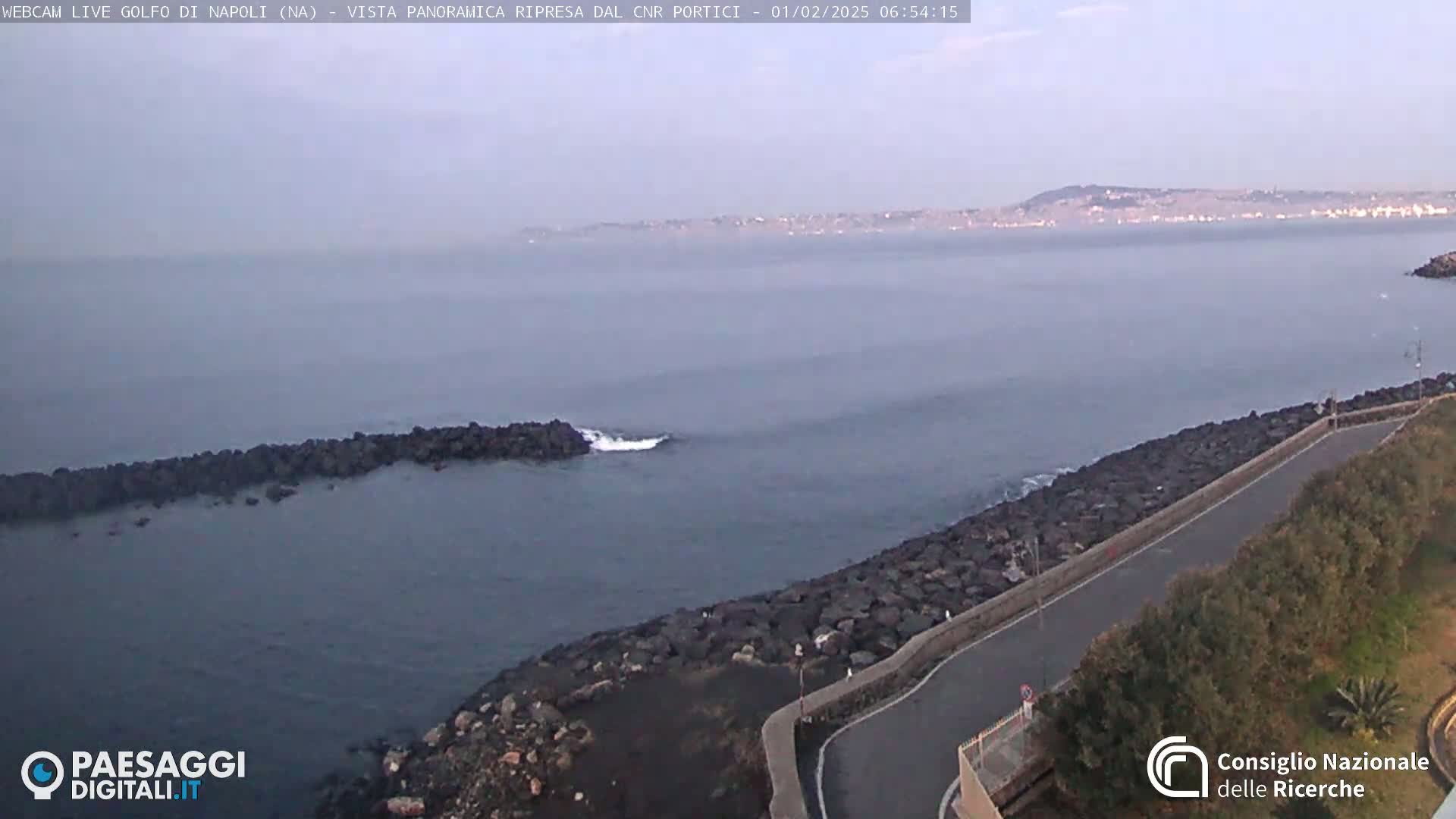 A hazy, early-morning view of a calm sea with a rocky shoreline and a road curving alongside it, a distant city skyline visible in the background.