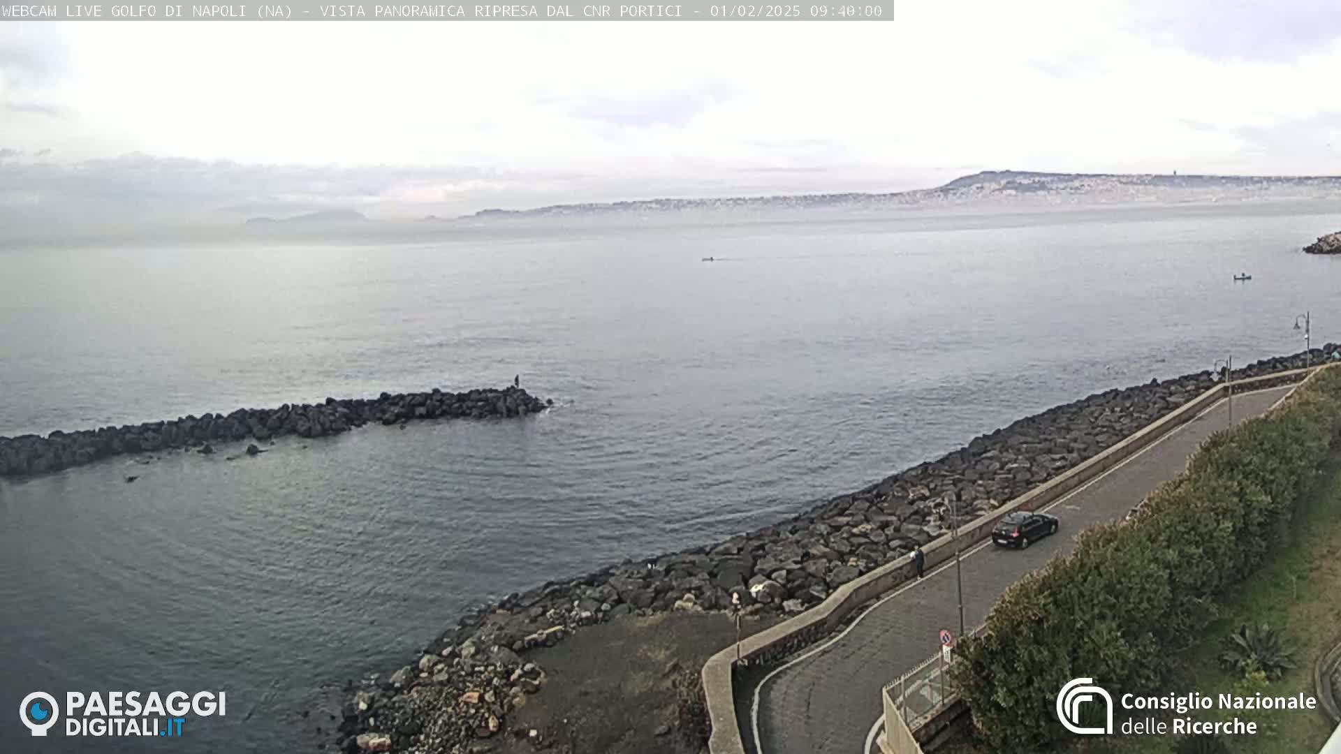 A rocky shoreline curves along a calm sea under an overcast sky, with a distant hazy mountain range visible.