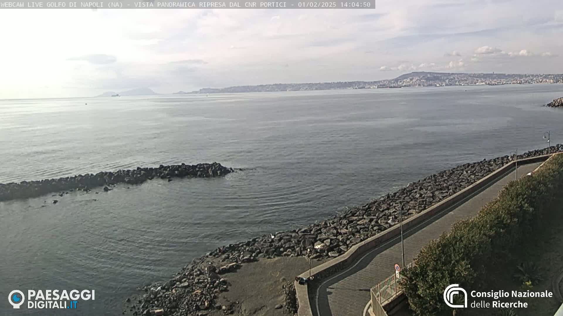 A rocky shoreline curves gently to the right, meeting a calm body of water that extends to a distant, hazy coastline under a mostly clear sky.