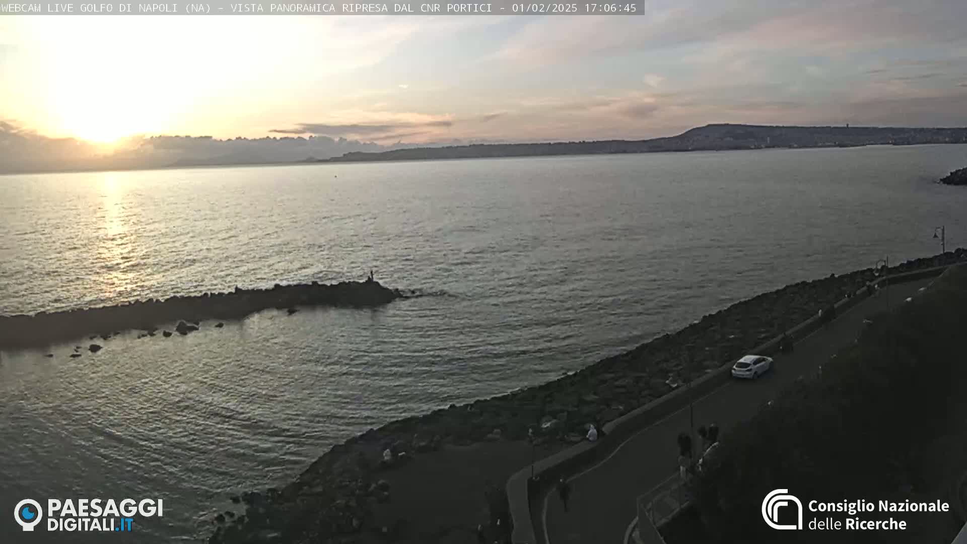 A calm sea at sunset is viewed from a rocky shoreline with a road and a few people visible, under a mostly clear sky.