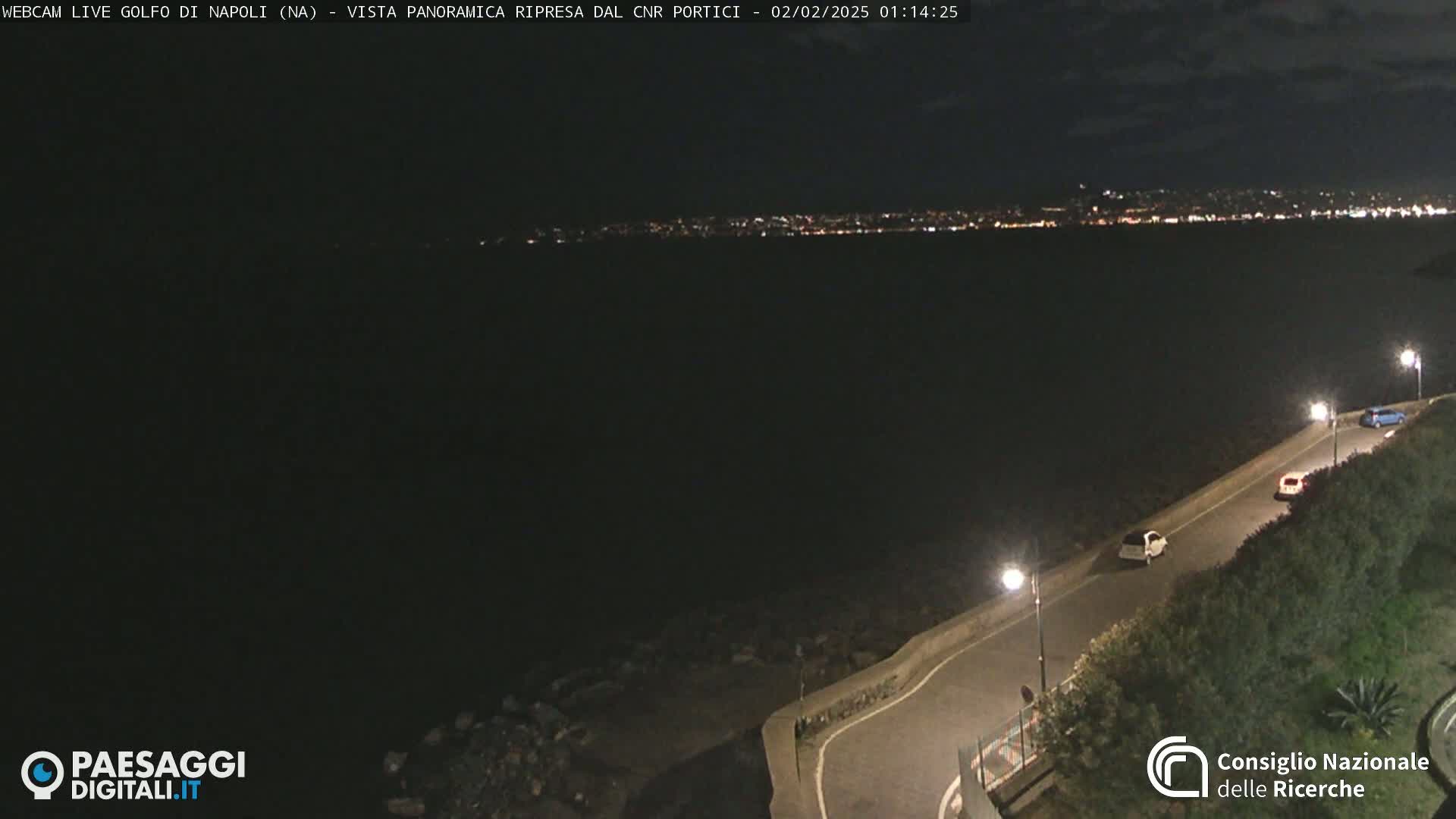 A nighttime, panoramic view of a dark body of water with a city's lights visible in the distance, a road curving along a shoreline with a few cars visible.