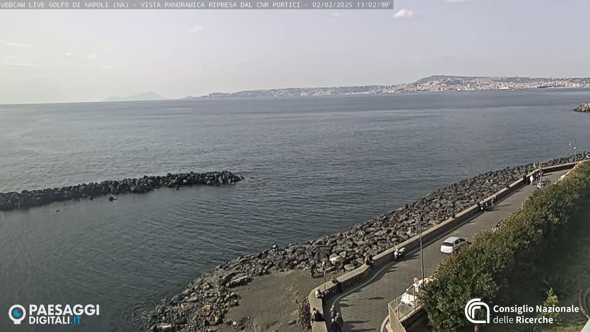 A rocky shoreline curves around a calm bay with a distant city and mountains visible under a mostly clear sky.
