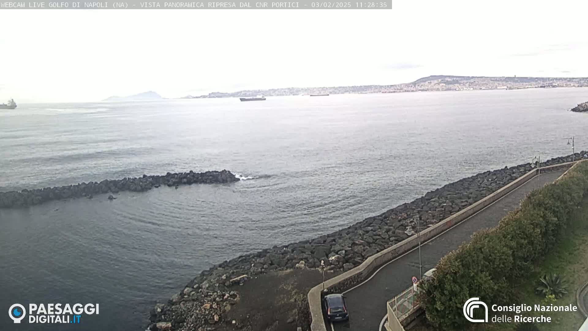 A rocky shoreline curves gently around a calm, gray body of water, with a distant hazy coastline and a few ships visible under an overcast sky.