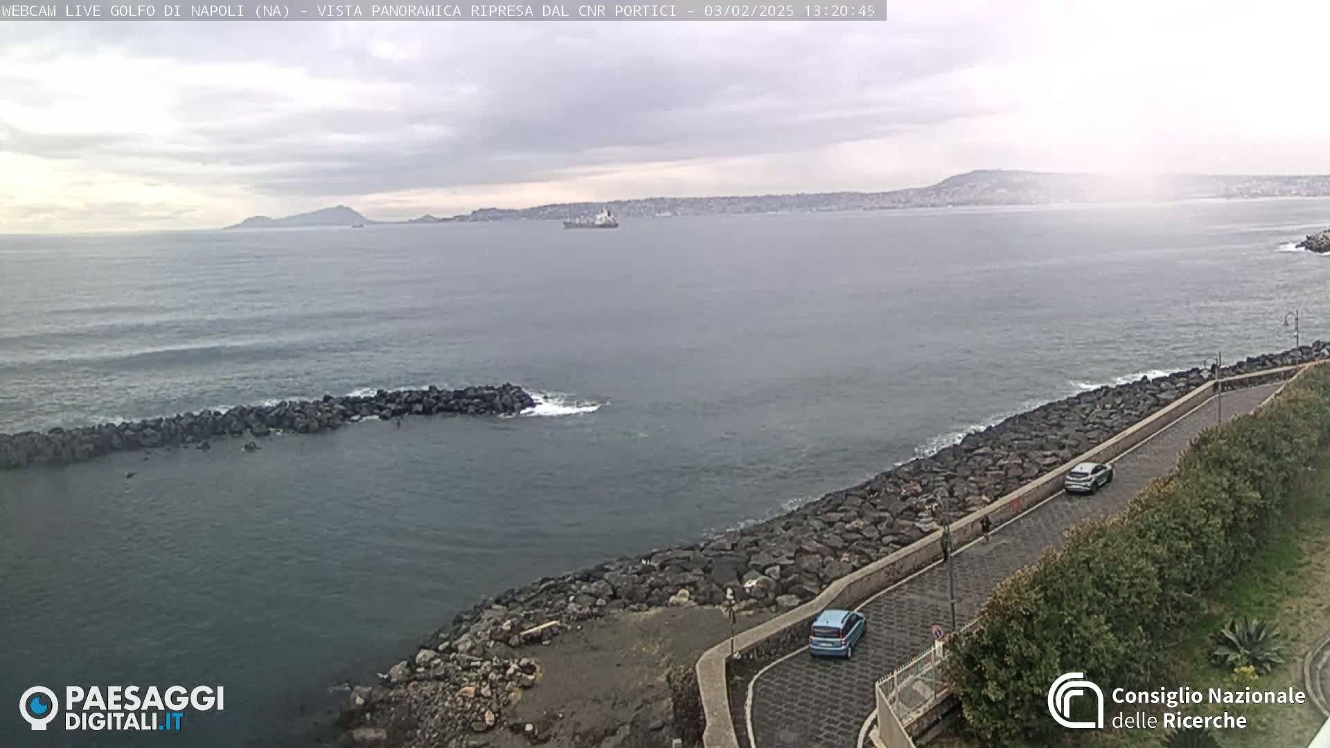 A coastal road curves along a rocky shoreline overlooking a calm sea under an overcast sky, with a distant mountain range visible on the horizon.