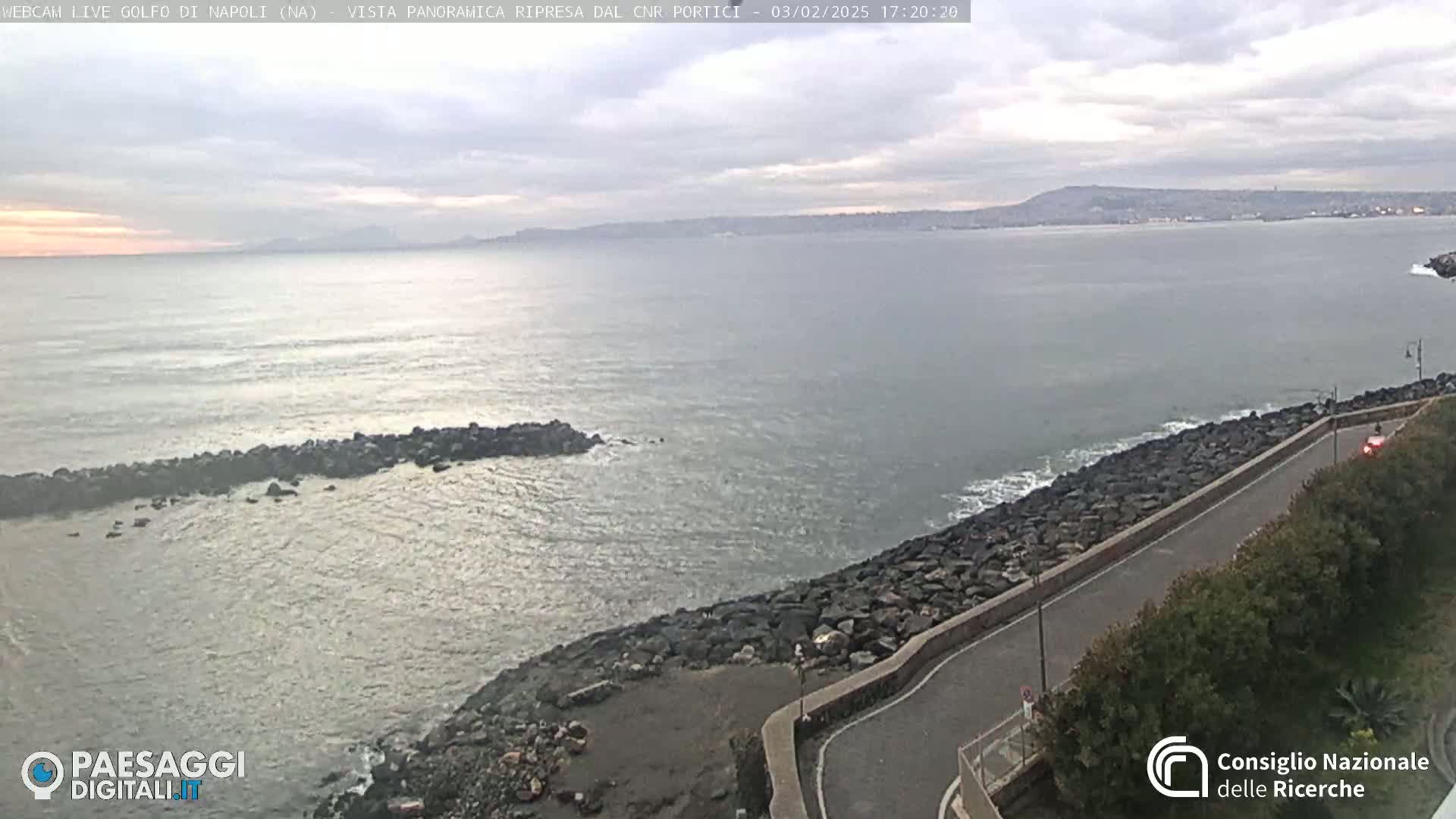 A rocky coastline with a road curves along the water's edge under a cloudy sky, overlooking a calm sea and distant mountains.
