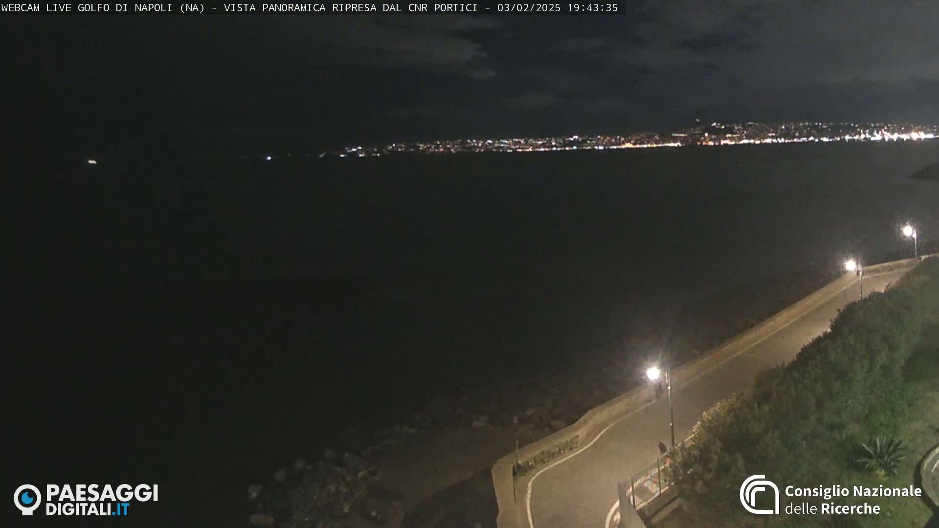 A nighttime, panoramic view of a dark body of water with a distant city skyline illuminated by lights, alongside a roadway curving along a shoreline.