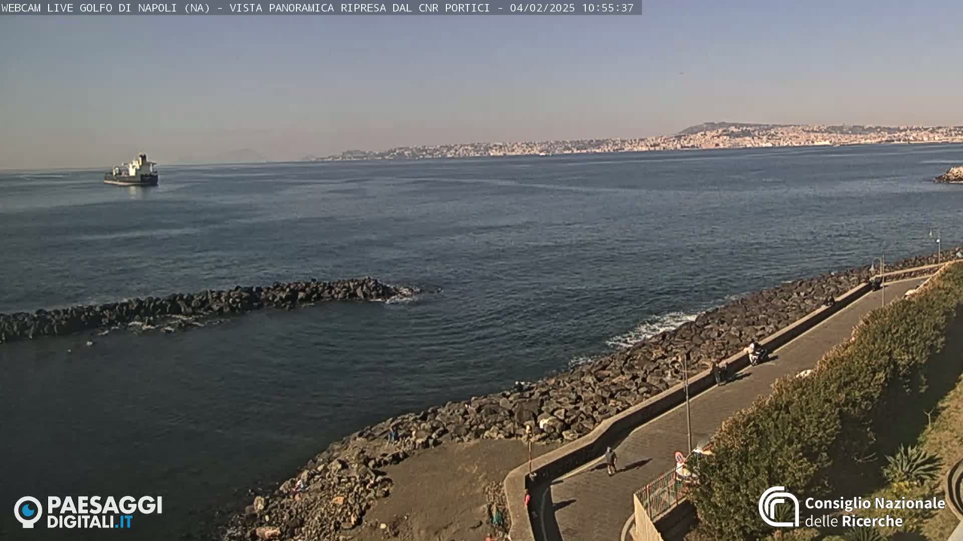 A cargo ship is visible on a calm, sunny sea, next to a rocky shoreline with a paved walkway and sparse vegetation.