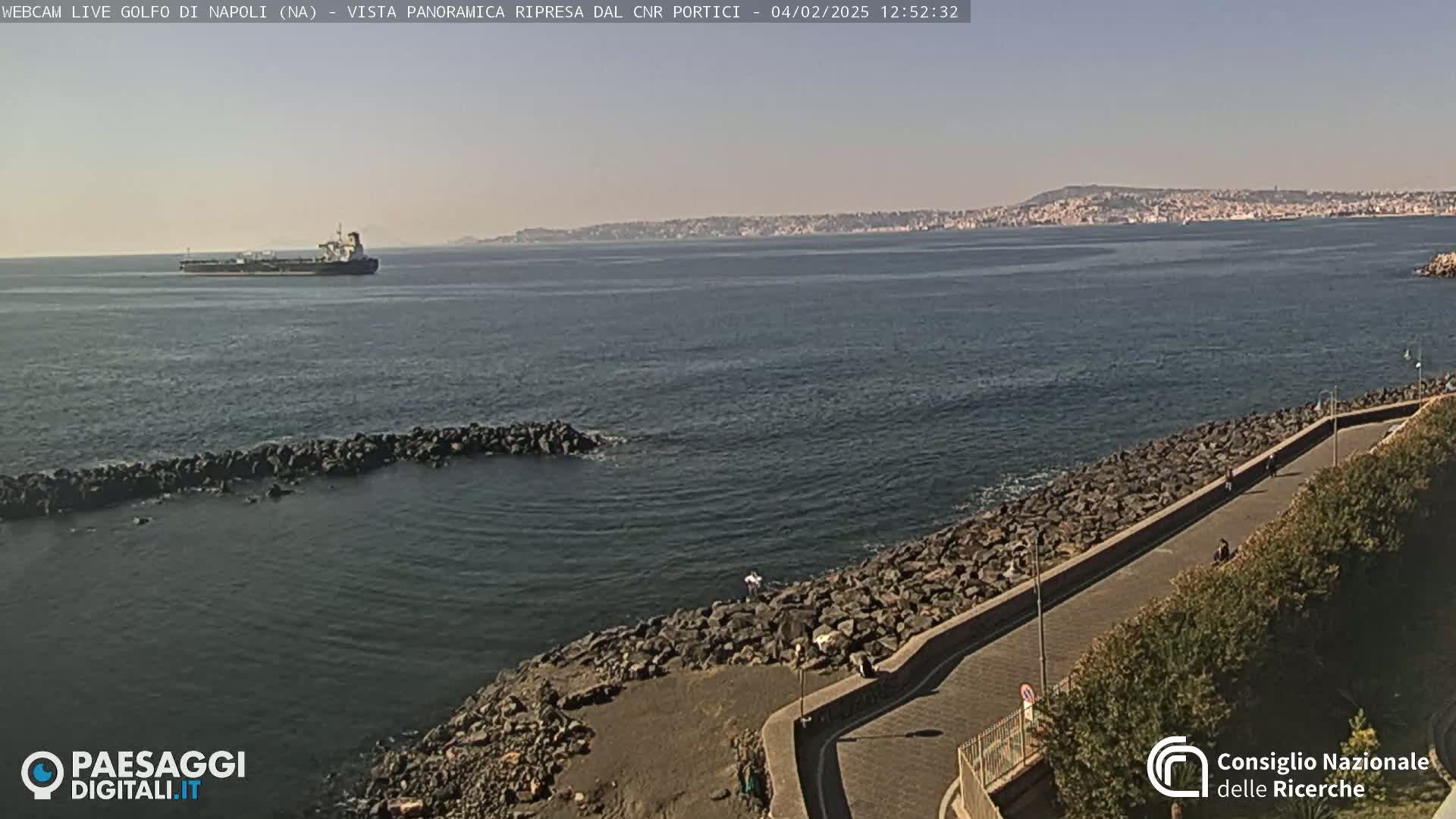 A large cargo ship is visible on a calm, sunny sea, near a rocky shoreline with a paved walkway and sparse vegetation.