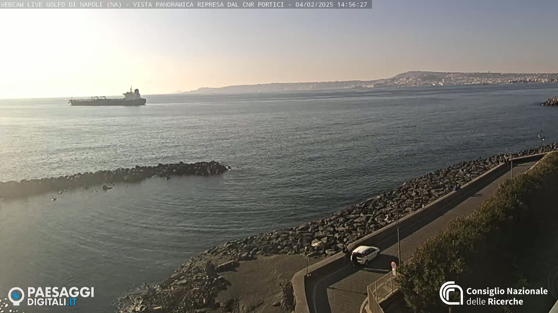 A large cargo ship is visible on a calm, clear sea near a rocky shoreline with a road and sparse vegetation; a city is visible in the distance.