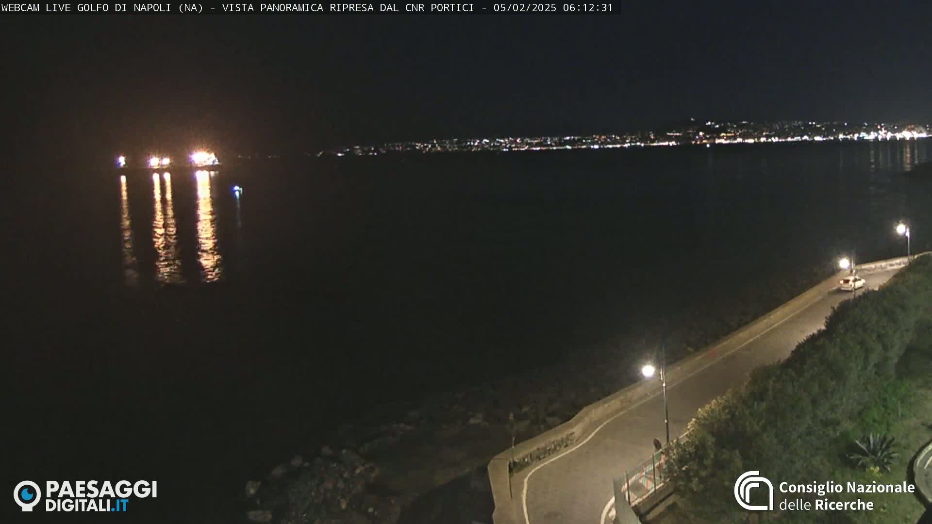 A dark nighttime scene shows a calm body of water reflecting the lights of a distant city, with a lone car driving along a coastal road lined with vegetation and illuminated by streetlights.
