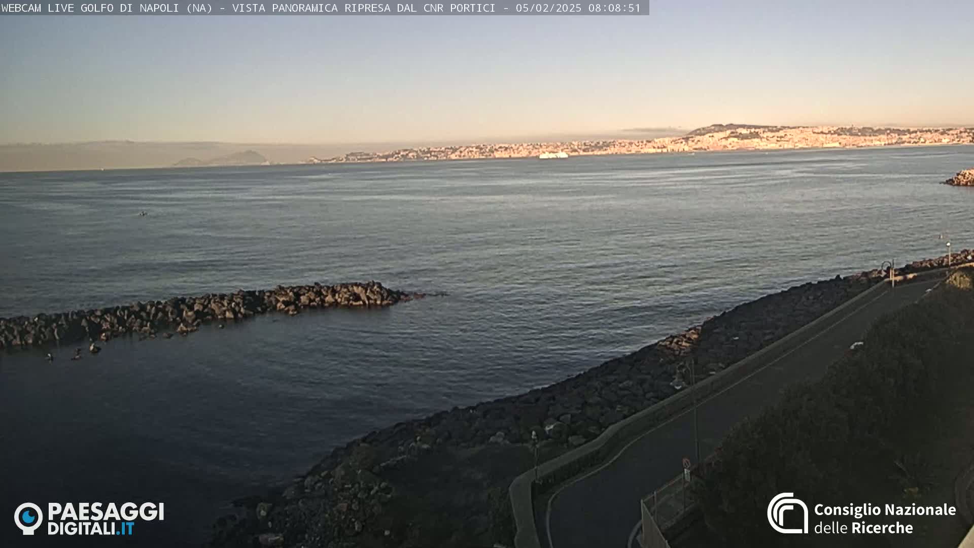 A panoramic view of a calm bay with a distant cityscape under a clear, sunrise sky shows a rocky shoreline and a paved road curving along the water's edge.