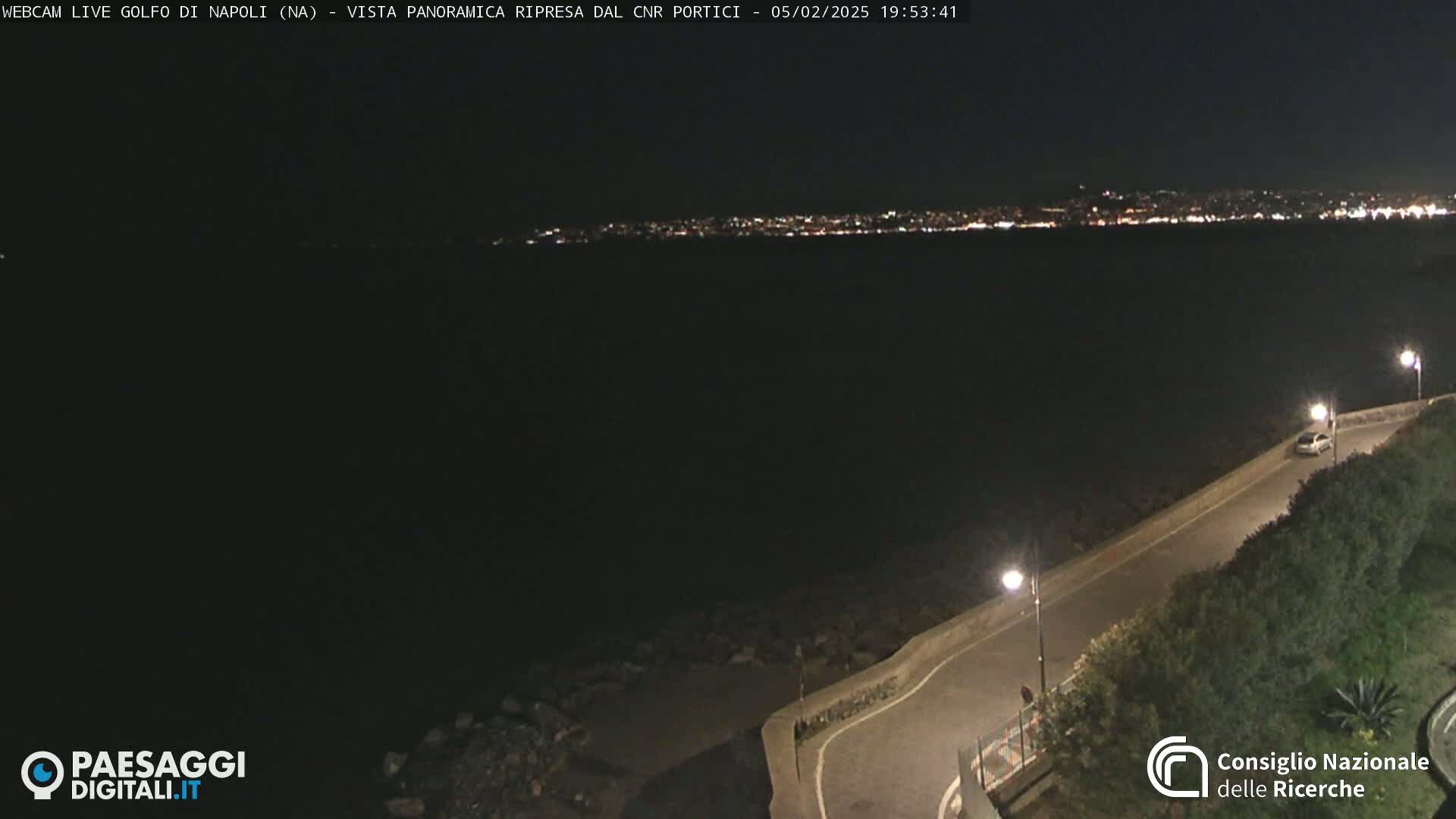 A nighttime, panoramic view shows a dark body of water with a distant, city skyline illuminated, and a curving road next to the water with a few lights and a lone car.