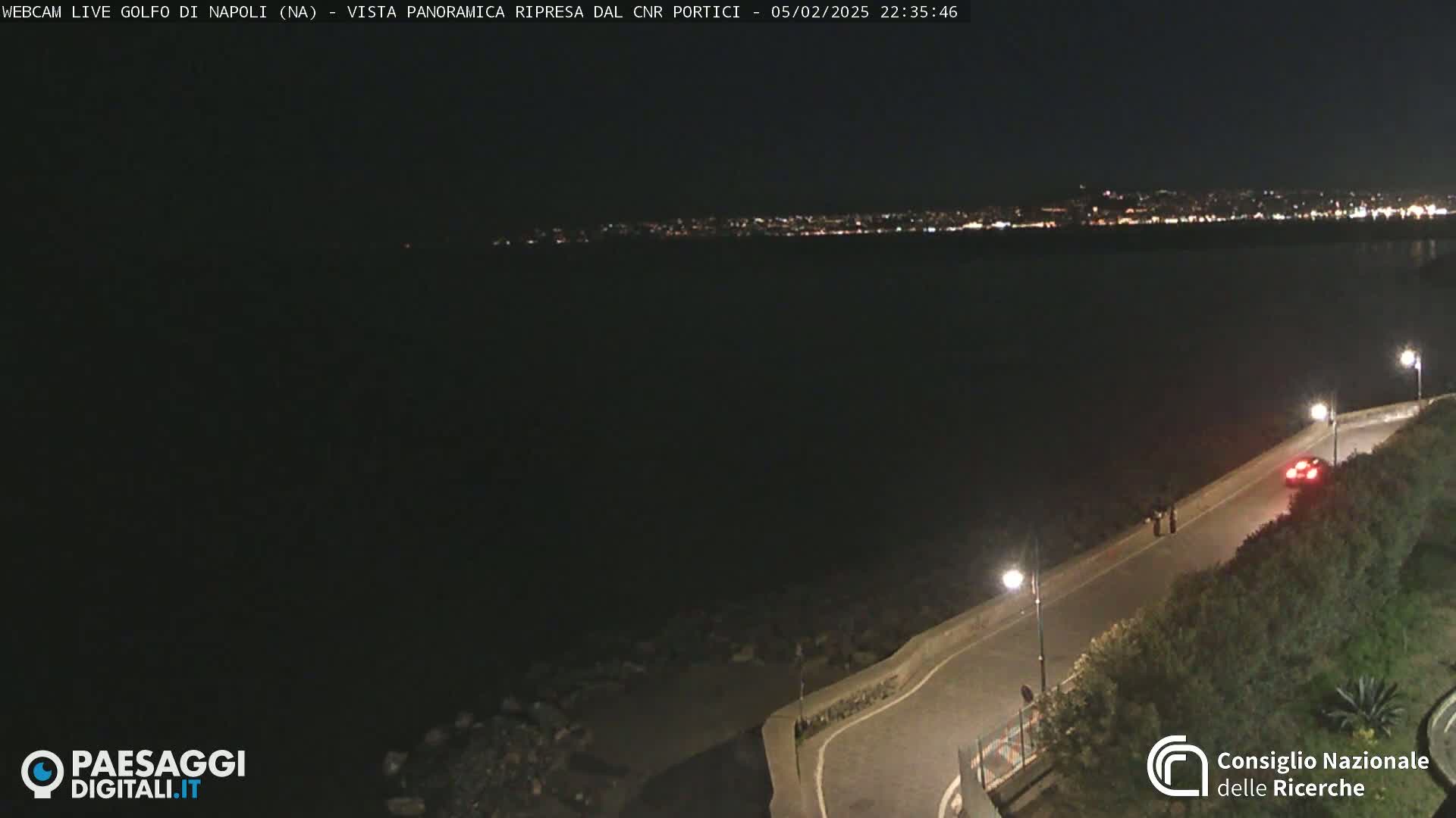 A nighttime, high-angle view shows a dark body of water reflecting the lights of a distant city across a curved road with sparse vegetation and a single car.