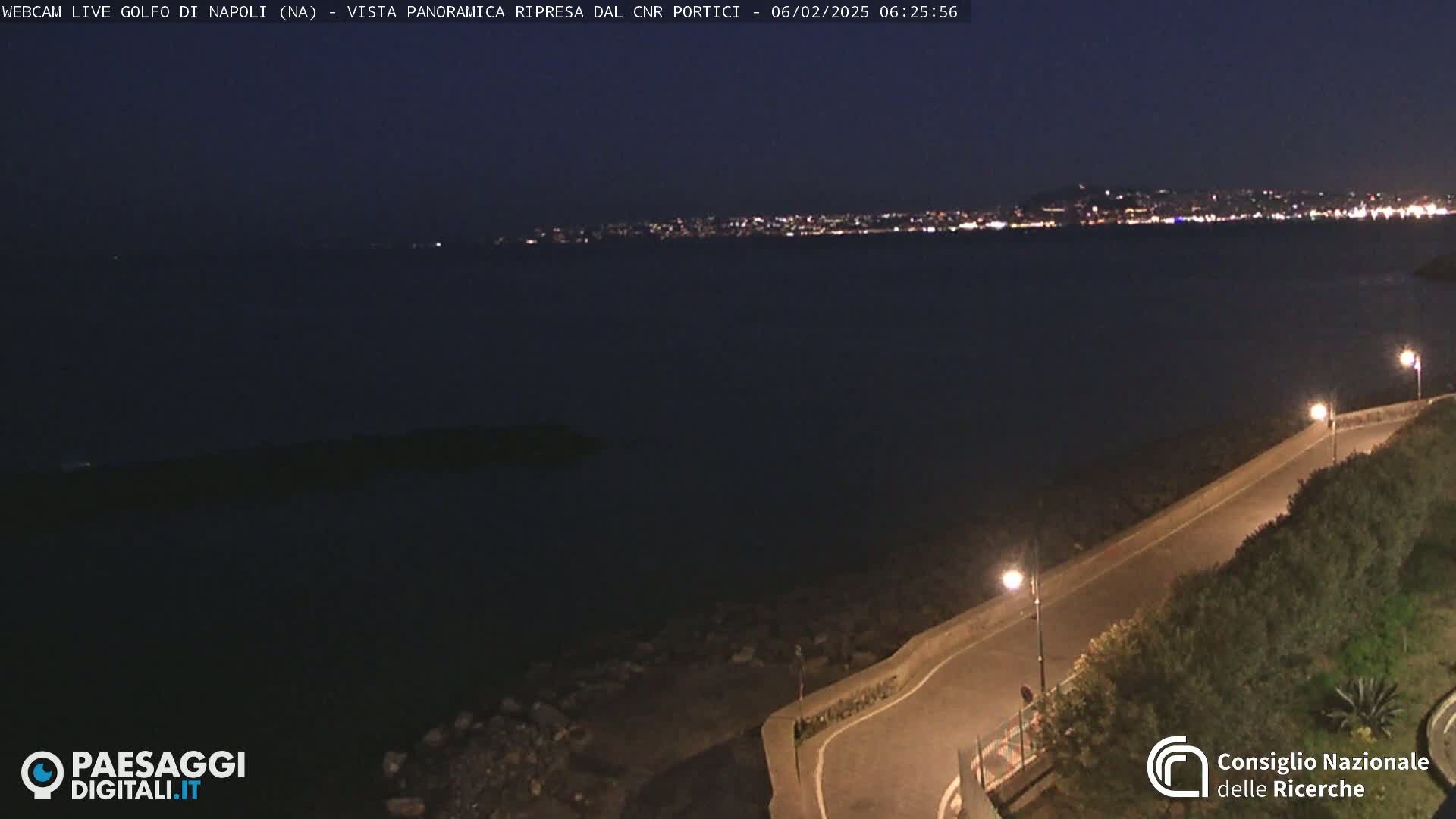 A nighttime, coastal view shows a dark body of water with a city skyline illuminated on the far shore, a curving road lit by streetlights alongside a rocky shoreline in the foreground.