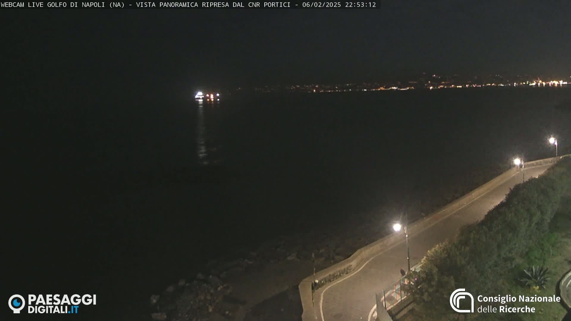 A nighttime, panoramic view shows a dark body of water with a distant city skyline illuminated by lights, a roadway curving along the water's edge, and a vessel in the water.